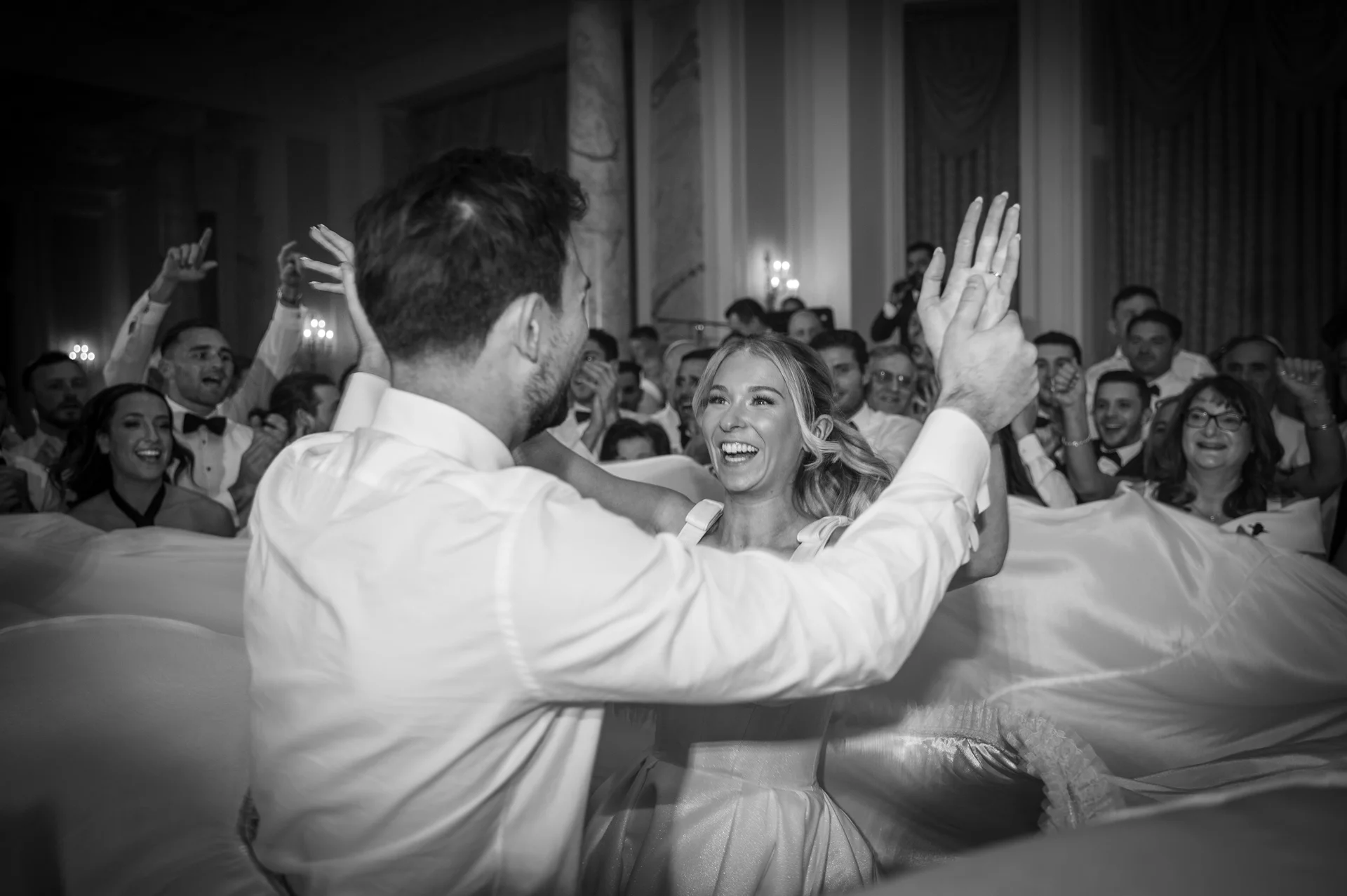 A joyful couple dance energetically together during their evening reception surrounded by a cheering crowd of wedding guests in a classic ballroom.