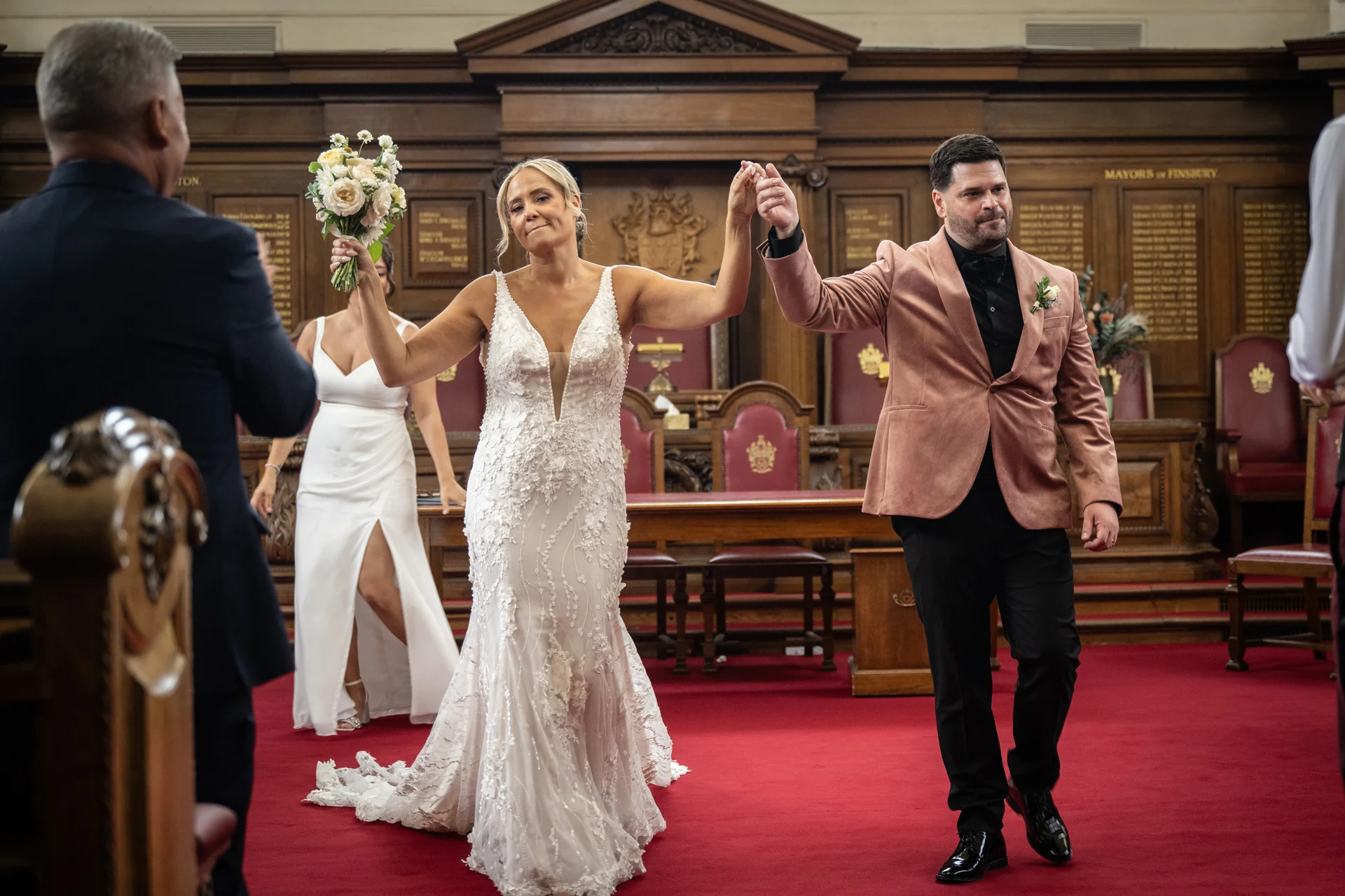 Newlyweds joyfully raise their joined hands as they walk down the aisle together after their town hall ceremony, with the bride holding a delicate bouquet.