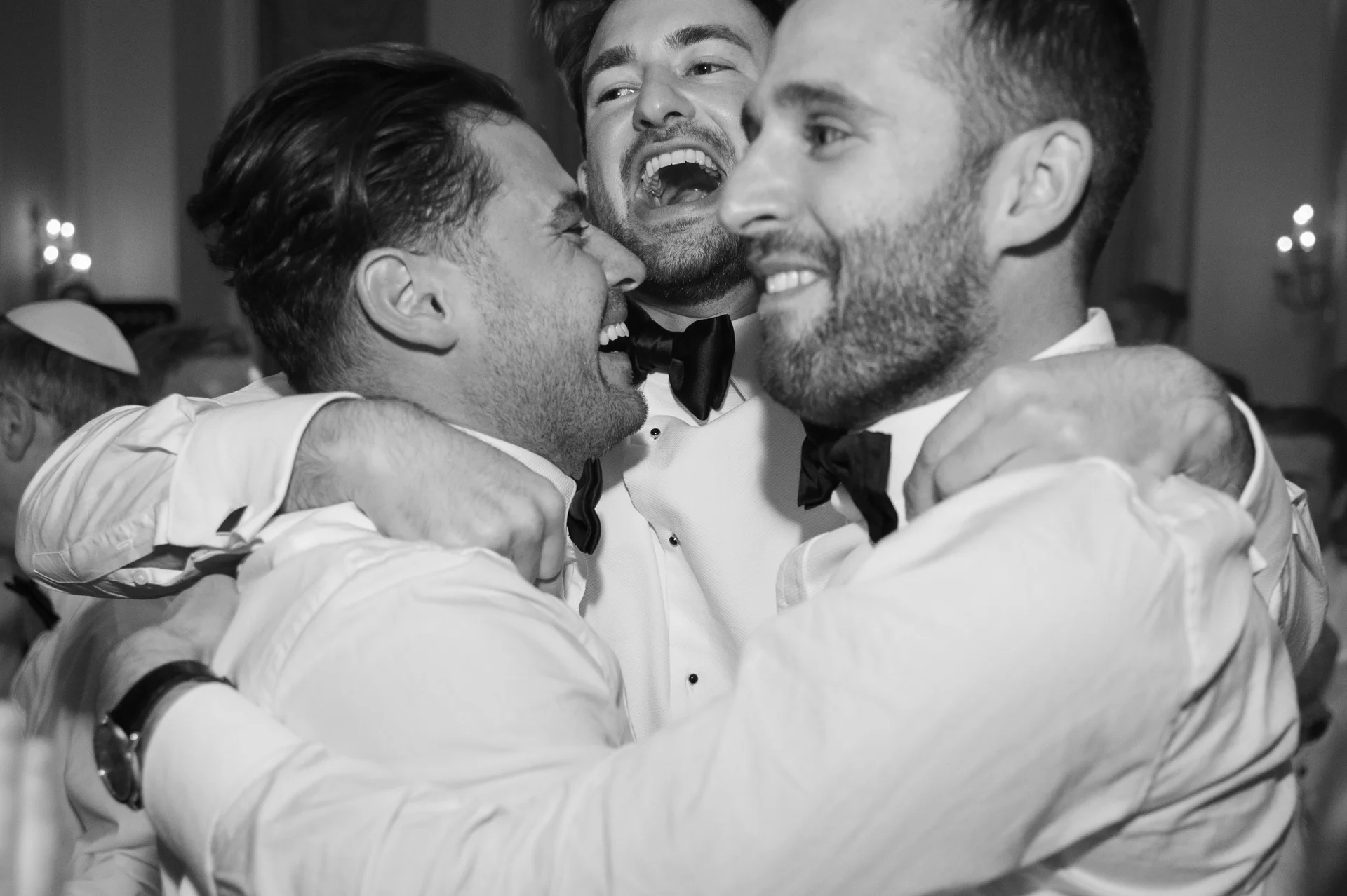 A group of groomsmen in black tie and white shirts share an exuberant, laughing embrace during the evening wedding reception.