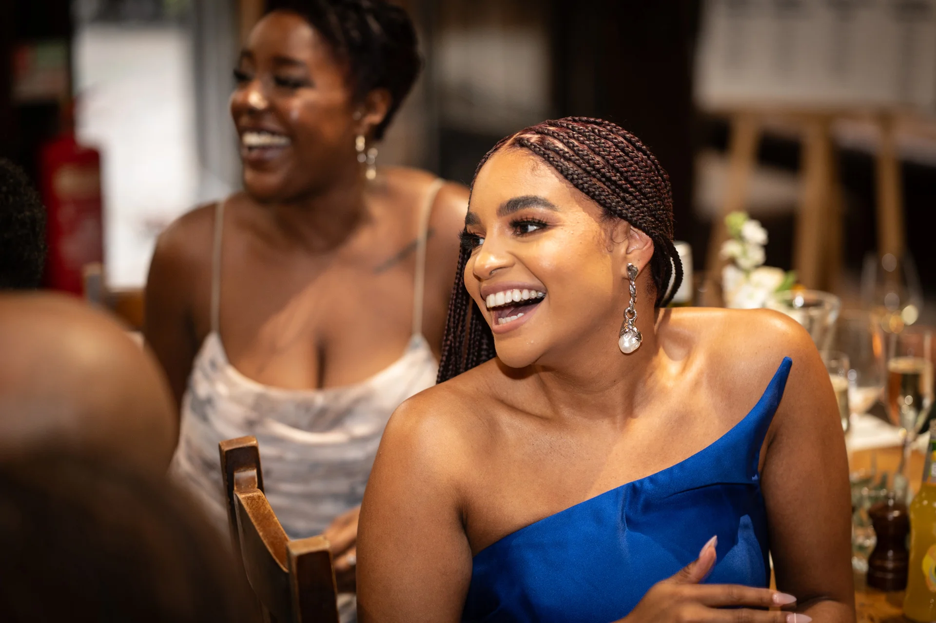 A smiling guest in a vibrant blue one-shoulder dress laughs joyfully during the wedding reception while seated at a table.