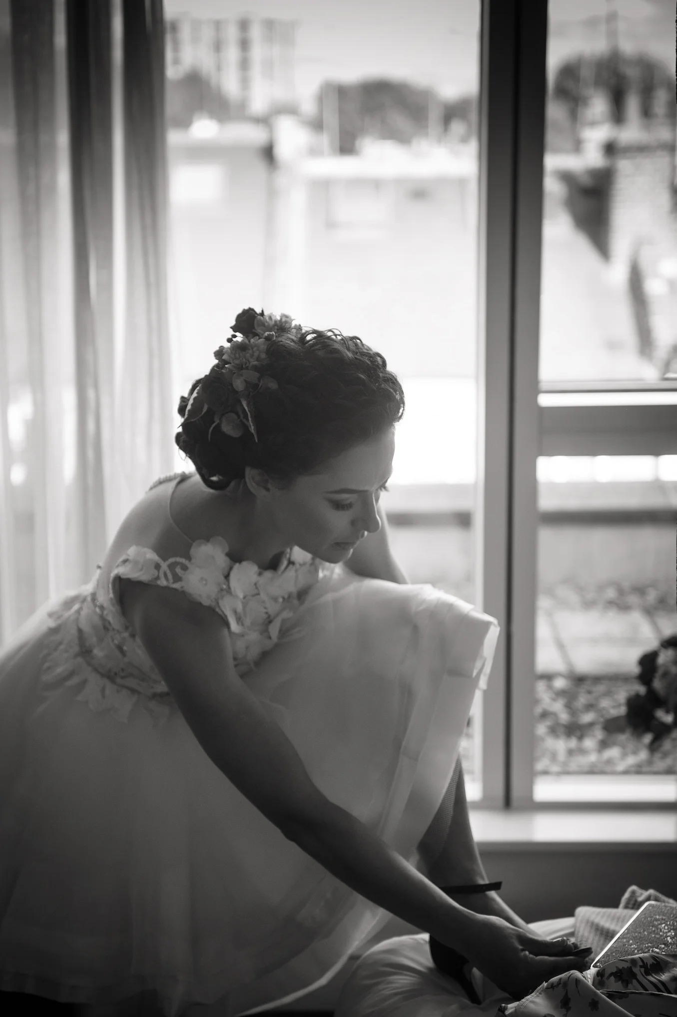 A black and white candid photograph of a bride putting on her wedding shoes near a bright window during bridal preparations.