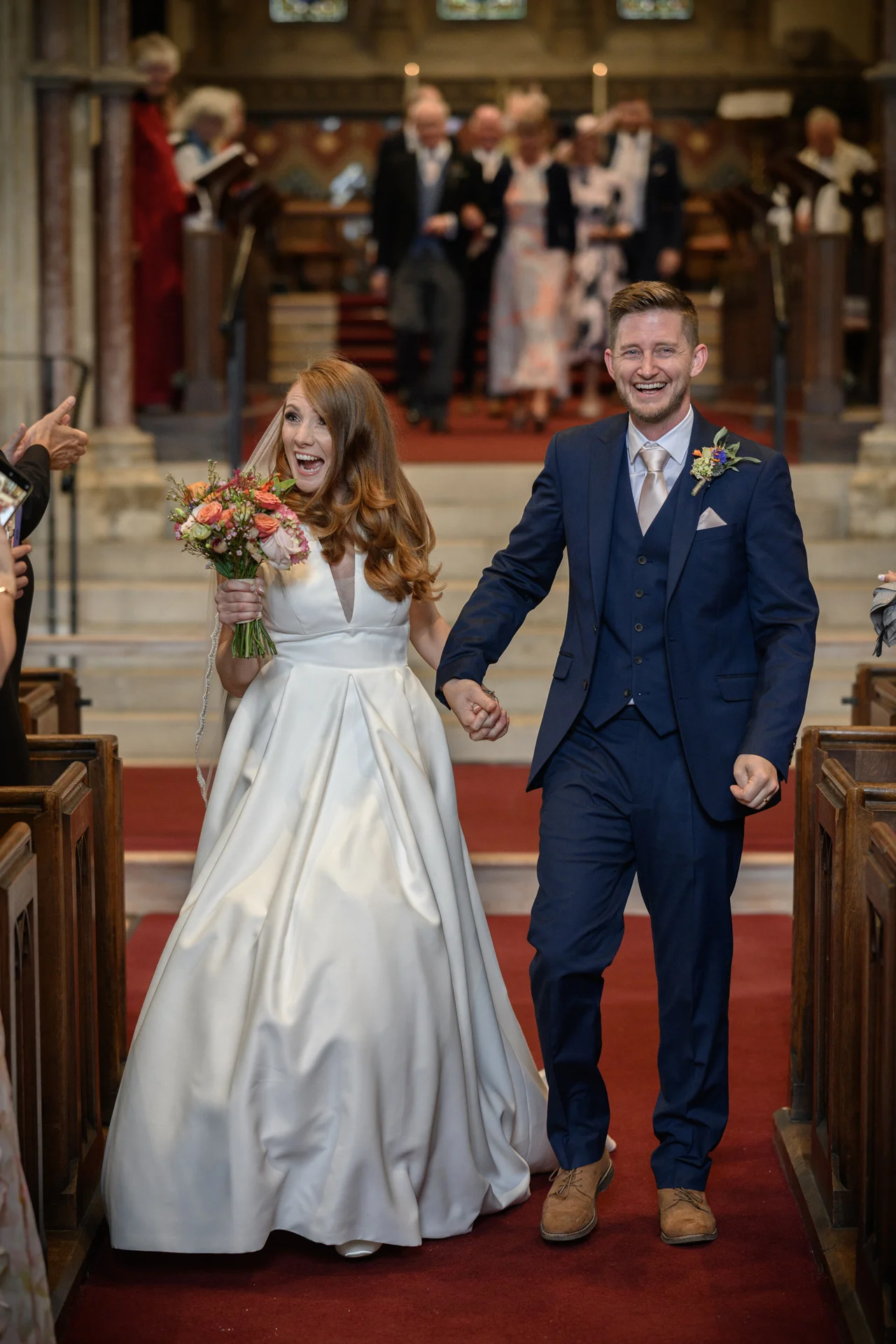 A joyous newlywed couple walks hand in hand down the aisle of a traditional church, laughing excitedly as guests cheer them on during the recessional.