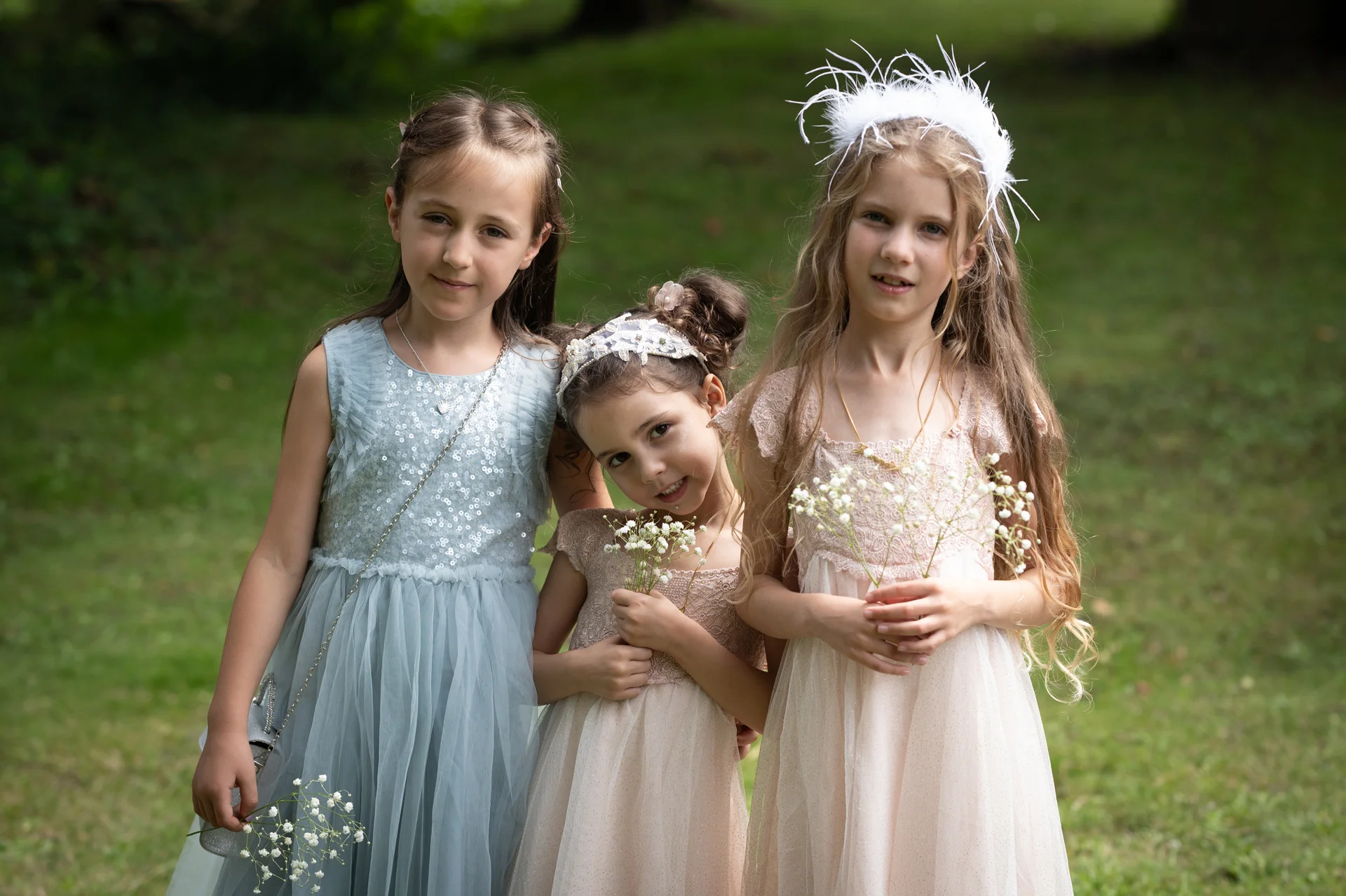 Three young flower girls and junior bridesmaids pose outdoors in the garden wearing pastel-coloured dresses and delicate hair accessories while holding small sprigs of gypsophila.