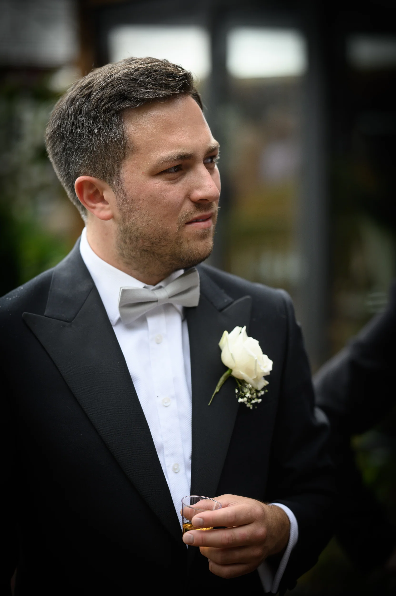 A handsome groom in a black tuxedo and grey bow tie holding a small glass of whiskey and wearing a white rose boutonniere.