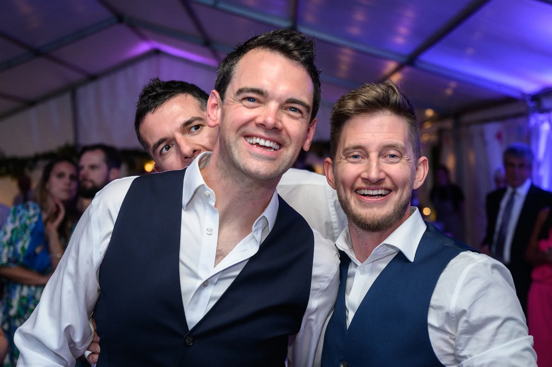 Three smiling men in waistcoats and white shirts pose together joyfully during a wedding reception illuminated by blue stage lighting inside a marquee.
