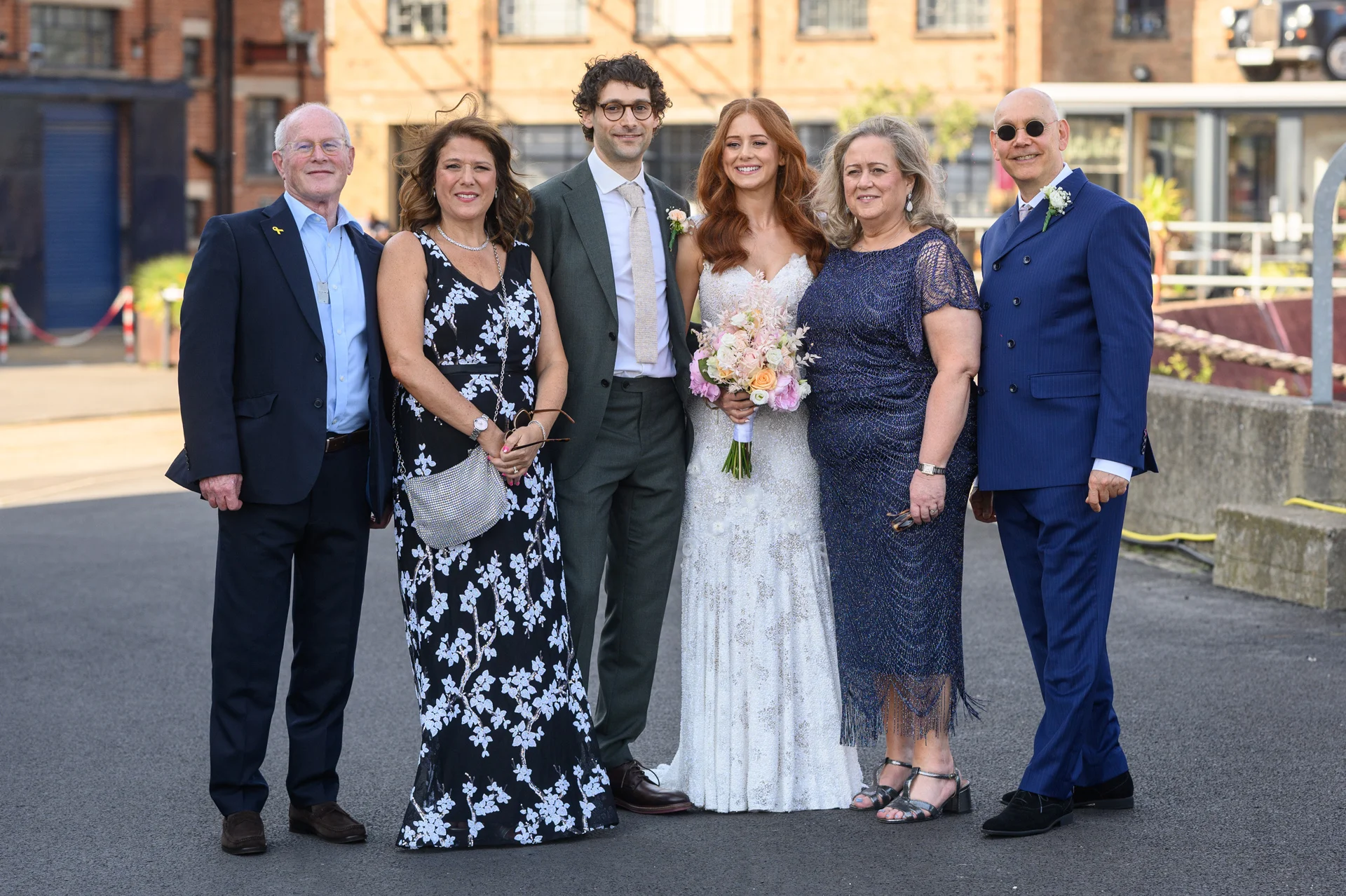 The bride and groom pose outdoors on a paved surface alongside family members, holding a delicate hand tied floral bouquet in natural daylight.