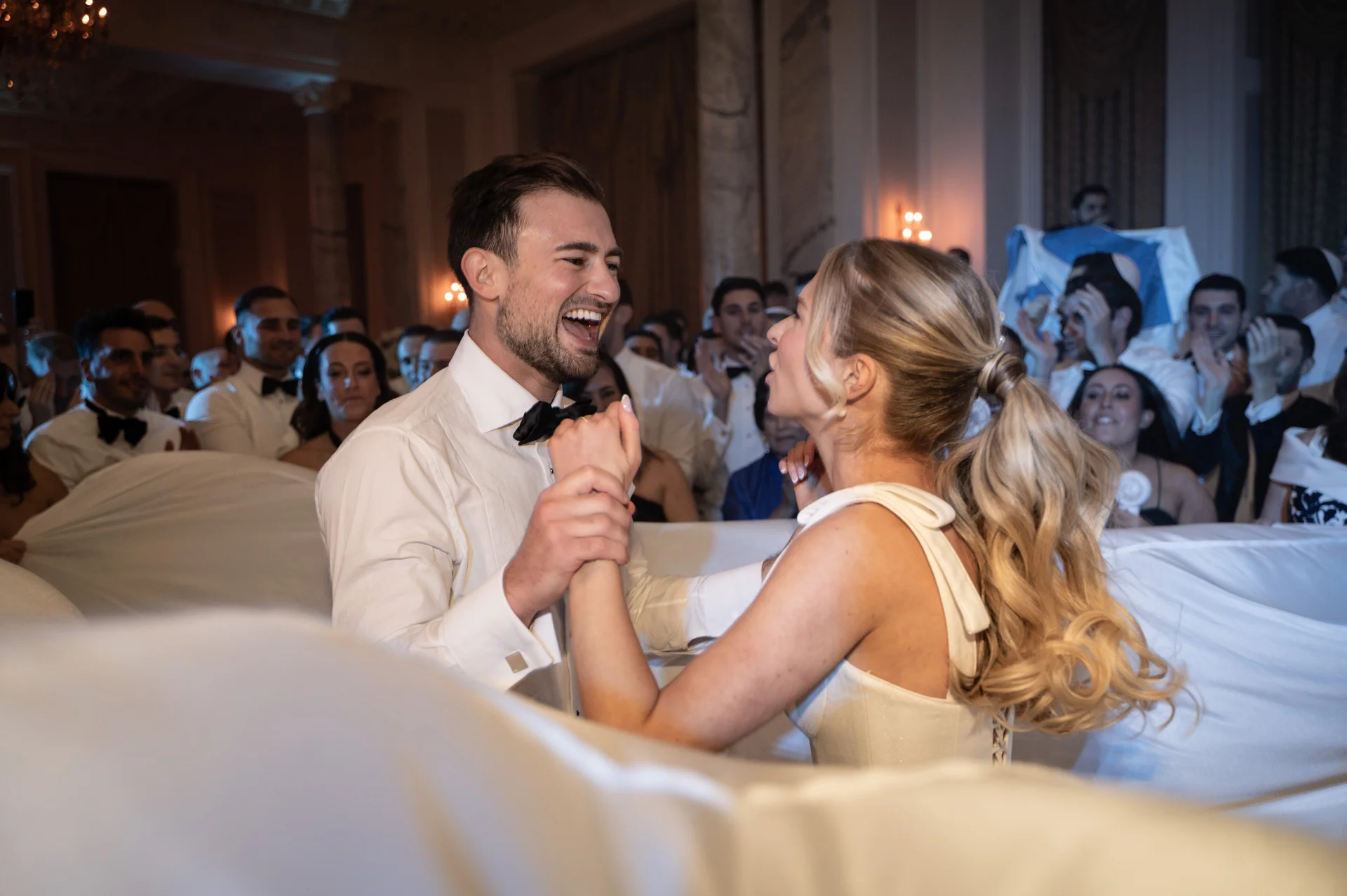 A joyous bride and groom laugh exuberantly while holding hands on the dance floor surrounded by cheering guests in a grand ballroom.