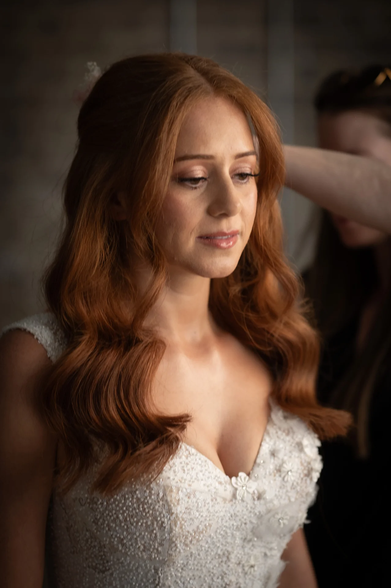 A redhead bride receives finishing touches to her half-up hair styling in a morning preparation moment, wearing a beaded sweetheart-neckline gown.