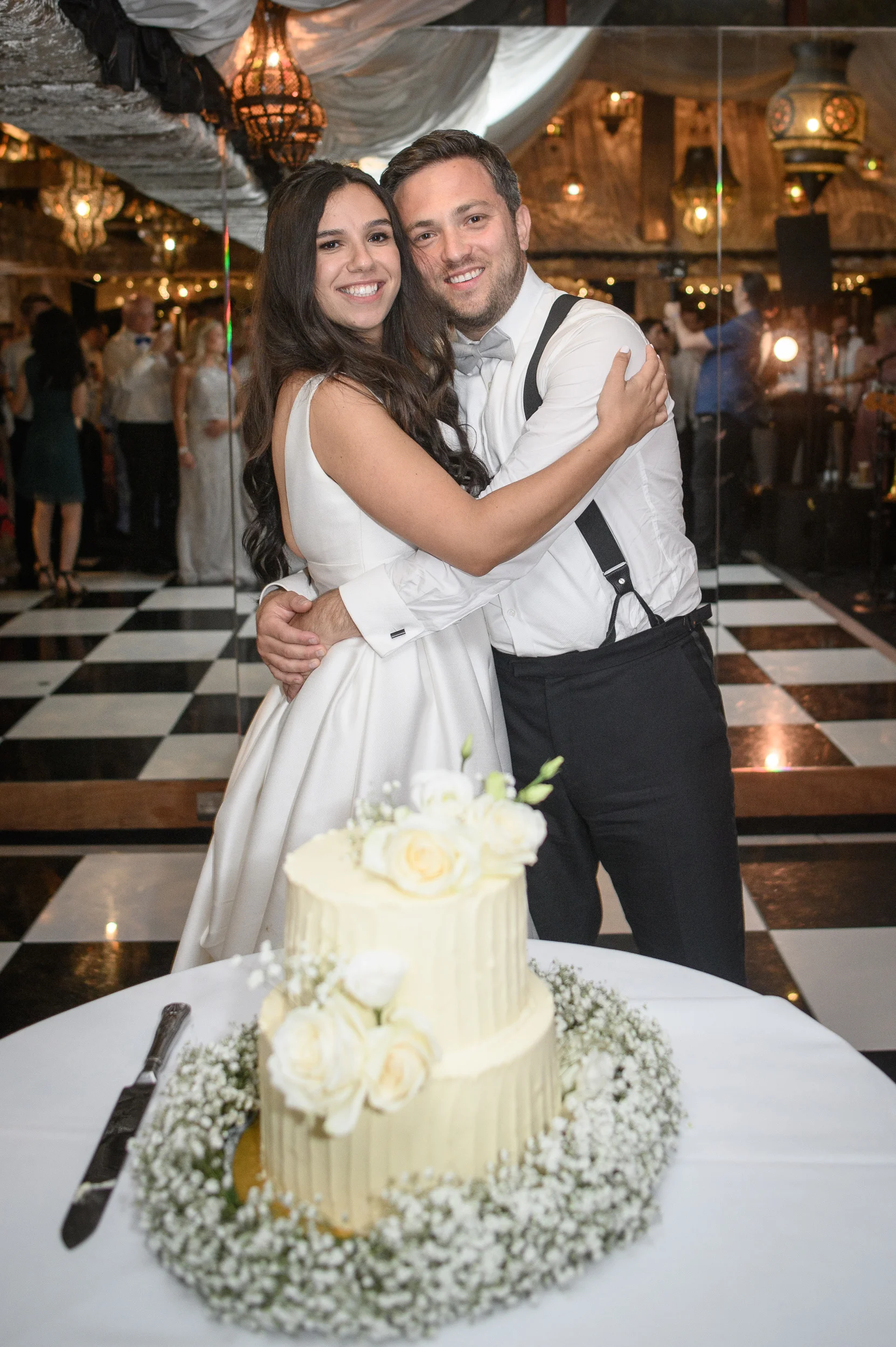A smiling bride and groom embrace warmly on a black and white checkerboard dance floor behind a two-tier white wedding cake decorated with roses.