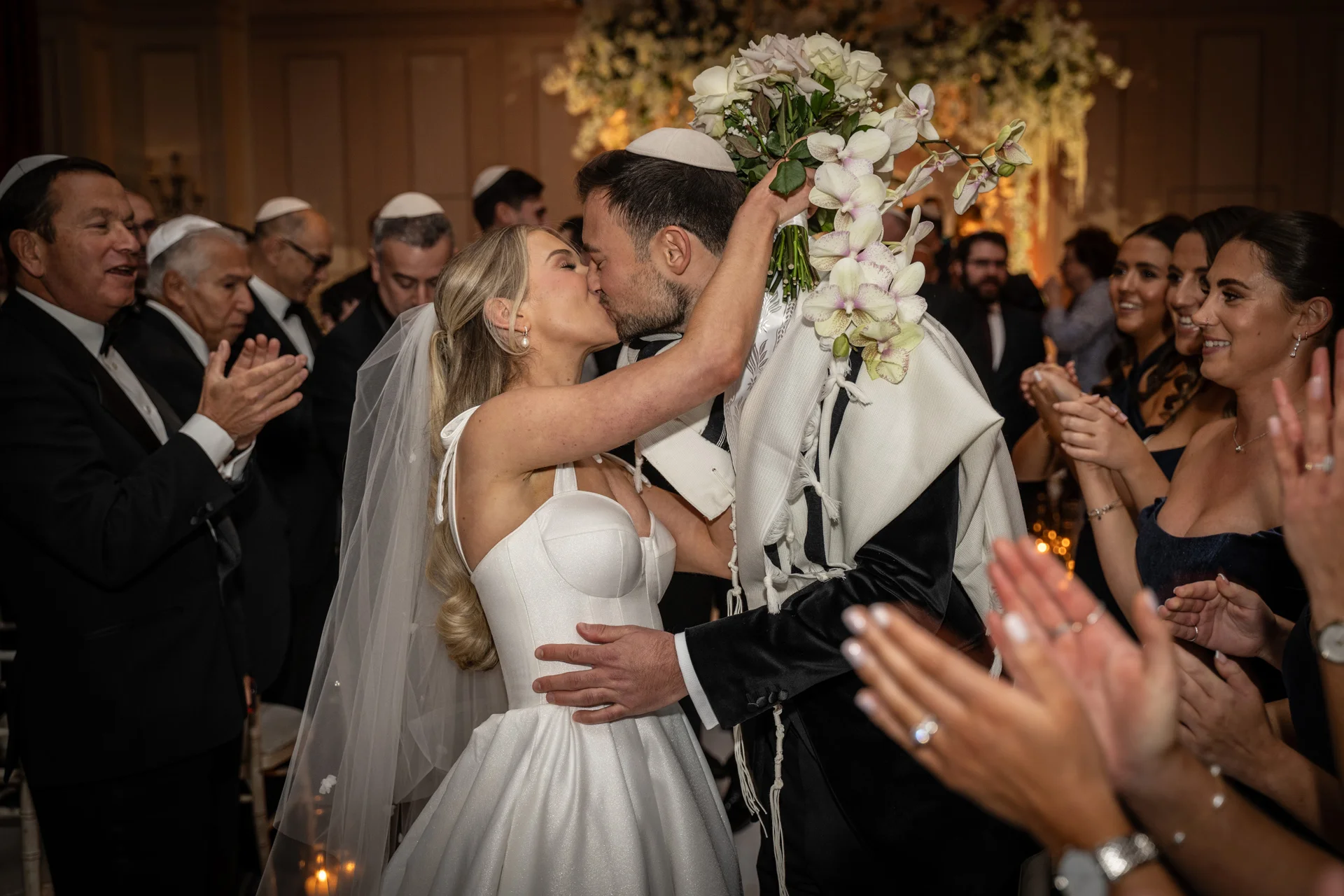 A newly married couple shares a passionate kiss during their Jewish wedding ceremony, surrounded by cheering guests as the groom wears a yarmulke and tallit while holding a cascading orchid bouquet.