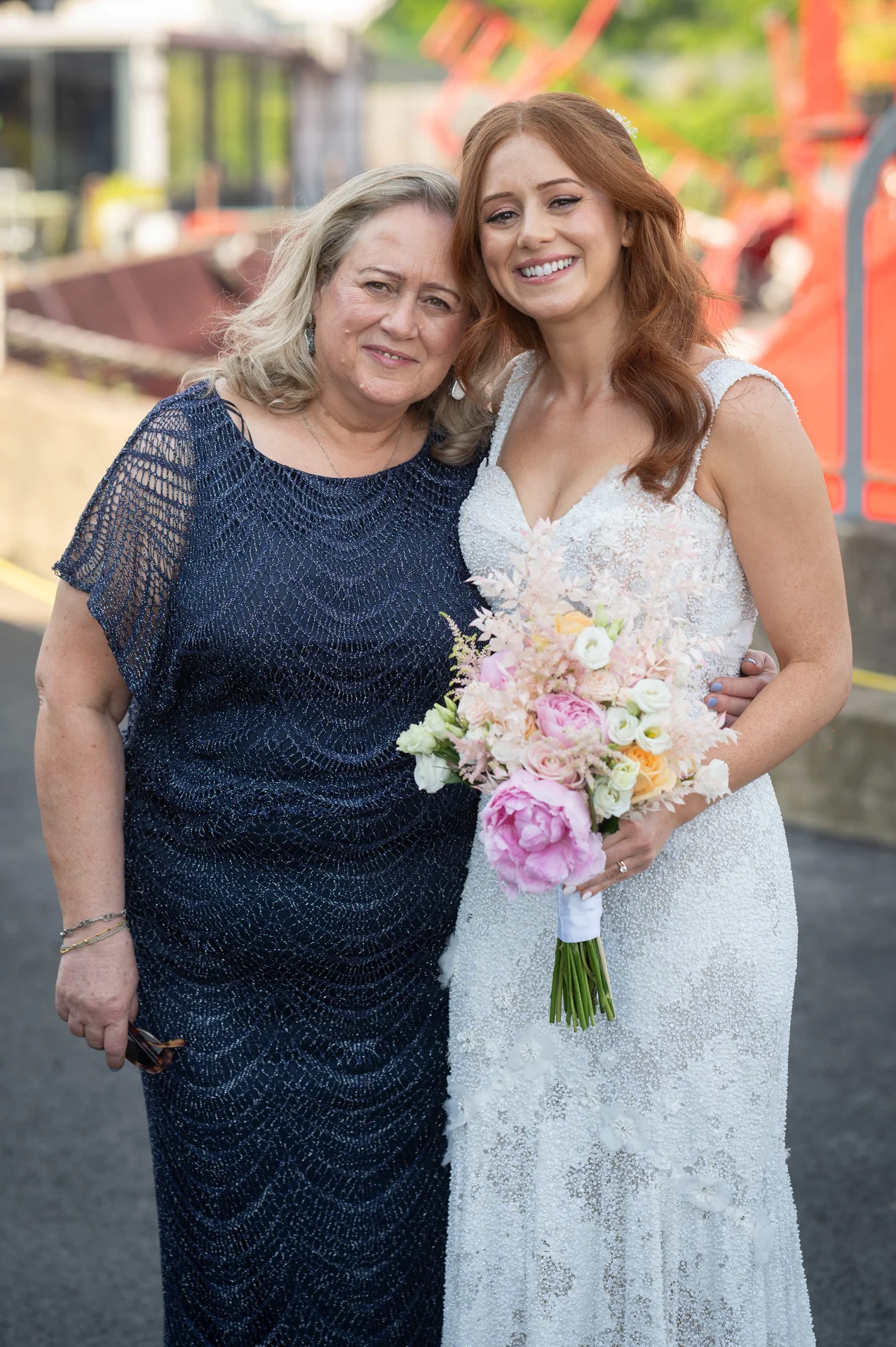A smiling red-haired bride in an embellished gown holding a pastel floral bouquet stands close to her mother, who is wearing a dark blue textured dress.