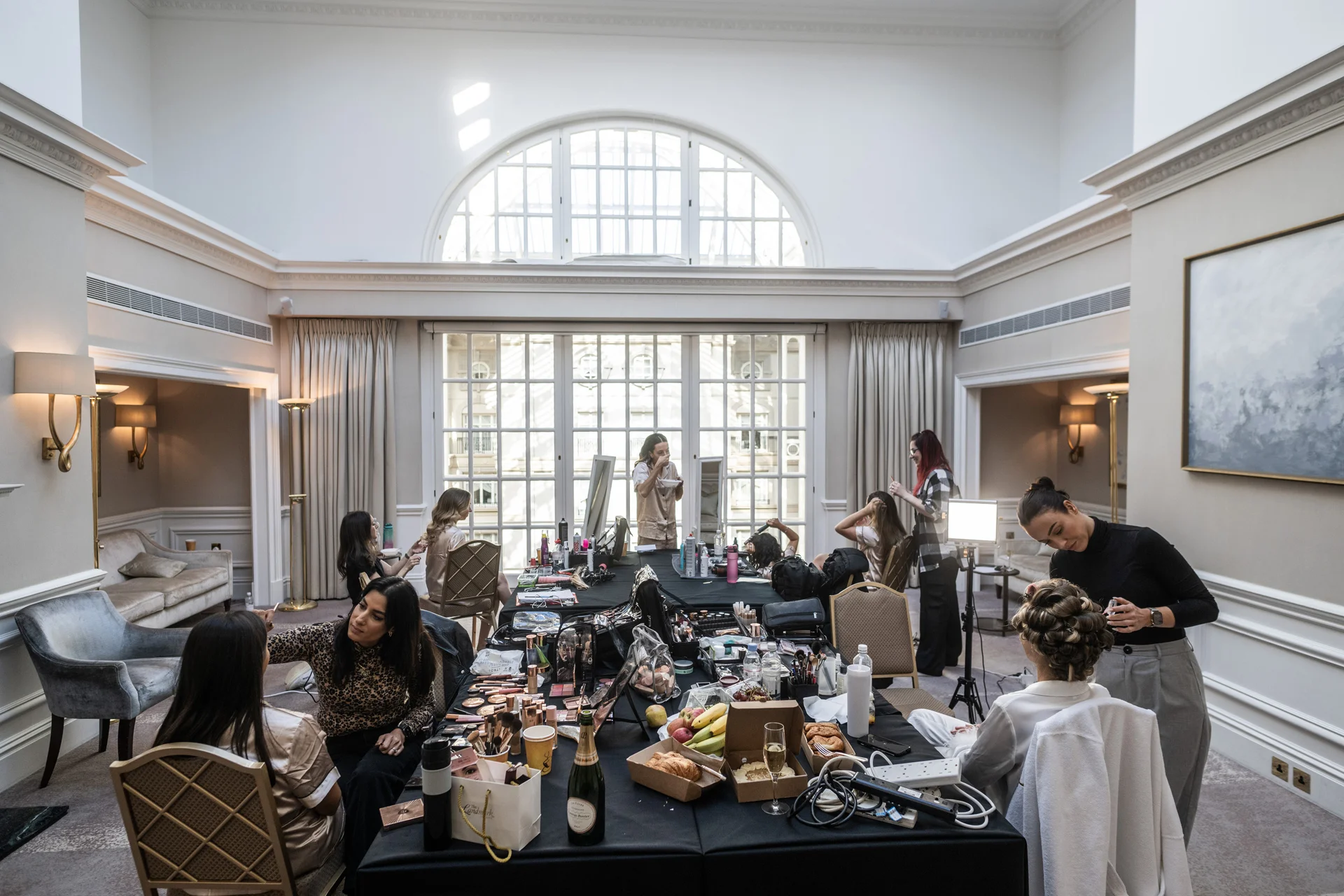 Bridal party and hair and makeup artists getting ready in a grand, sunlit ballroom with tall windows.