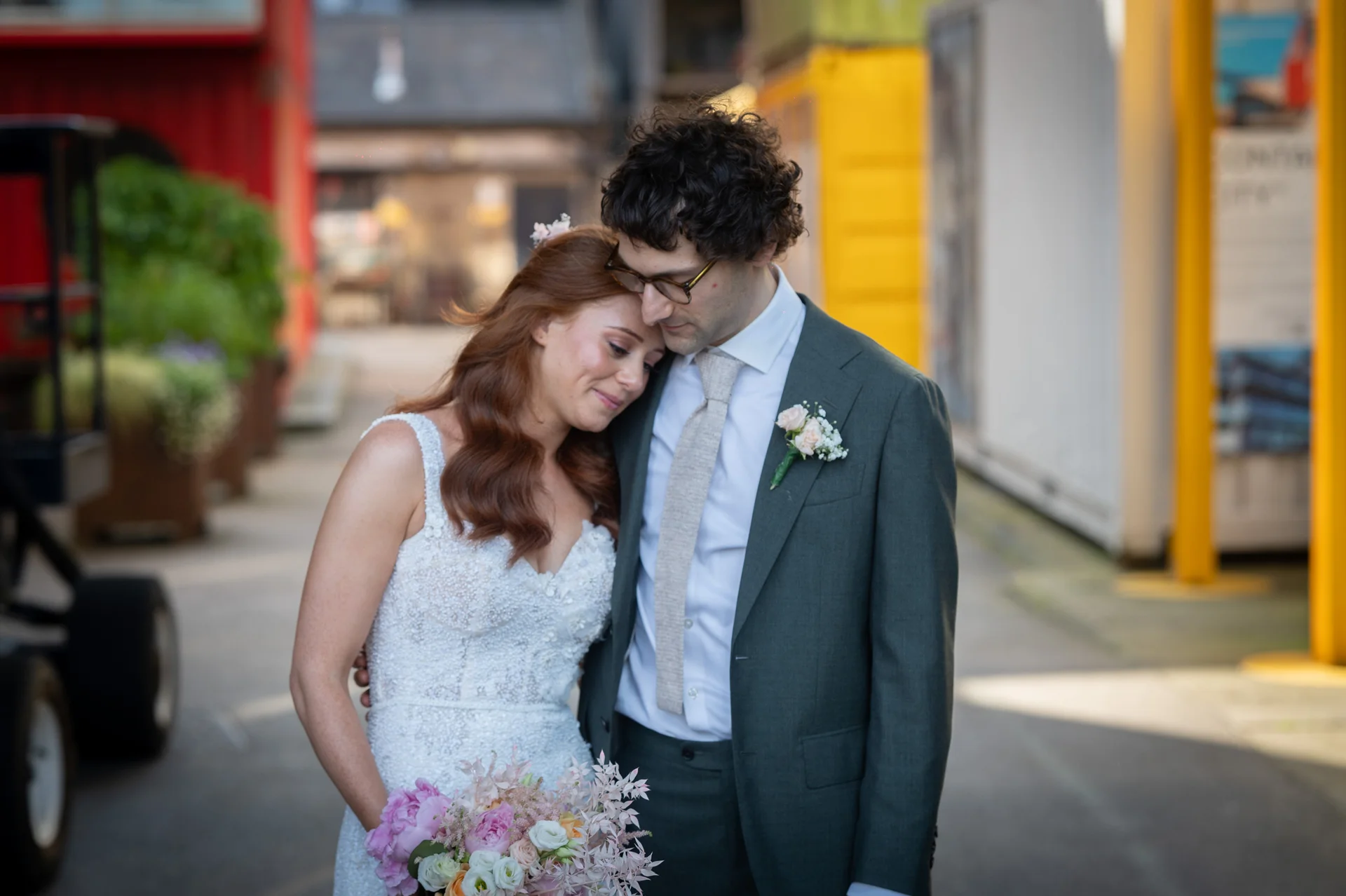 A newlywed couple shares a tender moment on an urban street, with the bride resting her head gently on the groom's shoulder.
