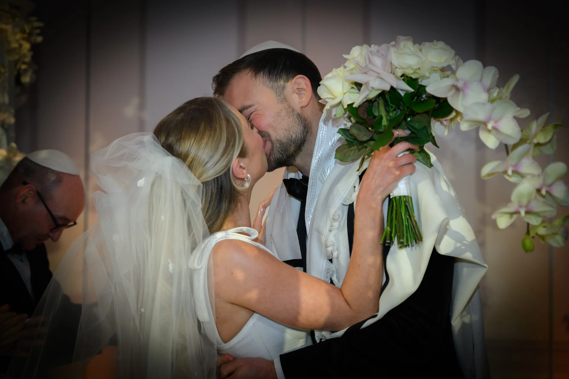 A newly married couple share a passionate first kiss during a traditional Jewish wedding ceremony, with the bride holding a lush bouquet of white roses and orchids.