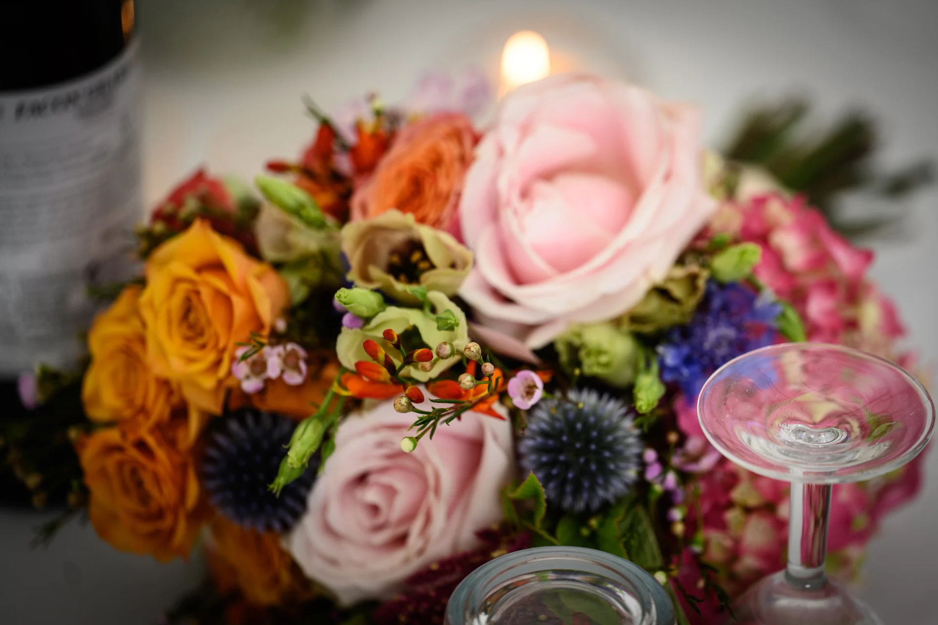 A vibrant wedding table arrangement featuring pink and orange roses, blue thistles, and delicate glassware illuminated by soft candlelight.