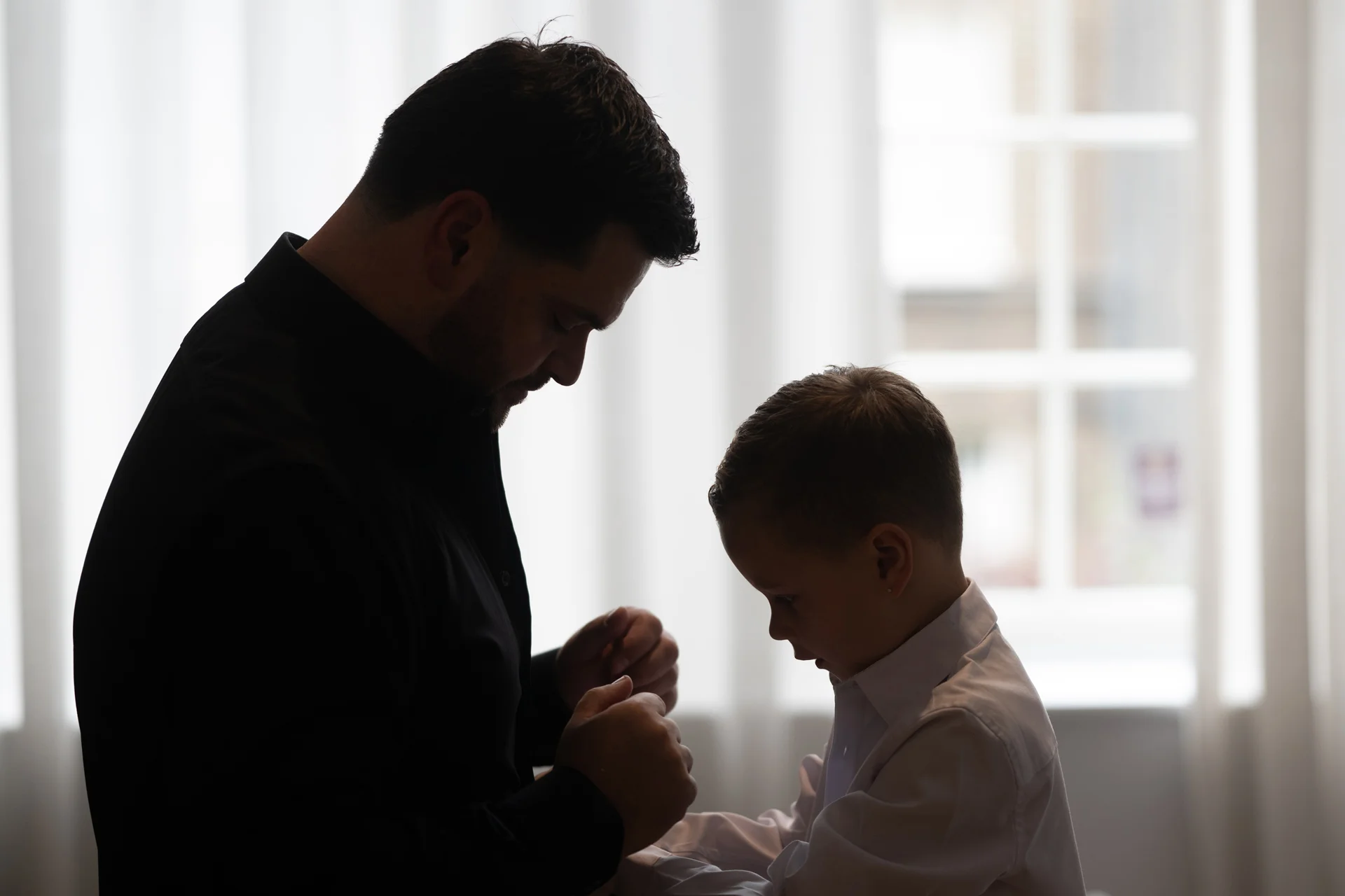 An adult groom and a young boy stand against a brightly lit window, captured in a quiet moment during wedding preparations.