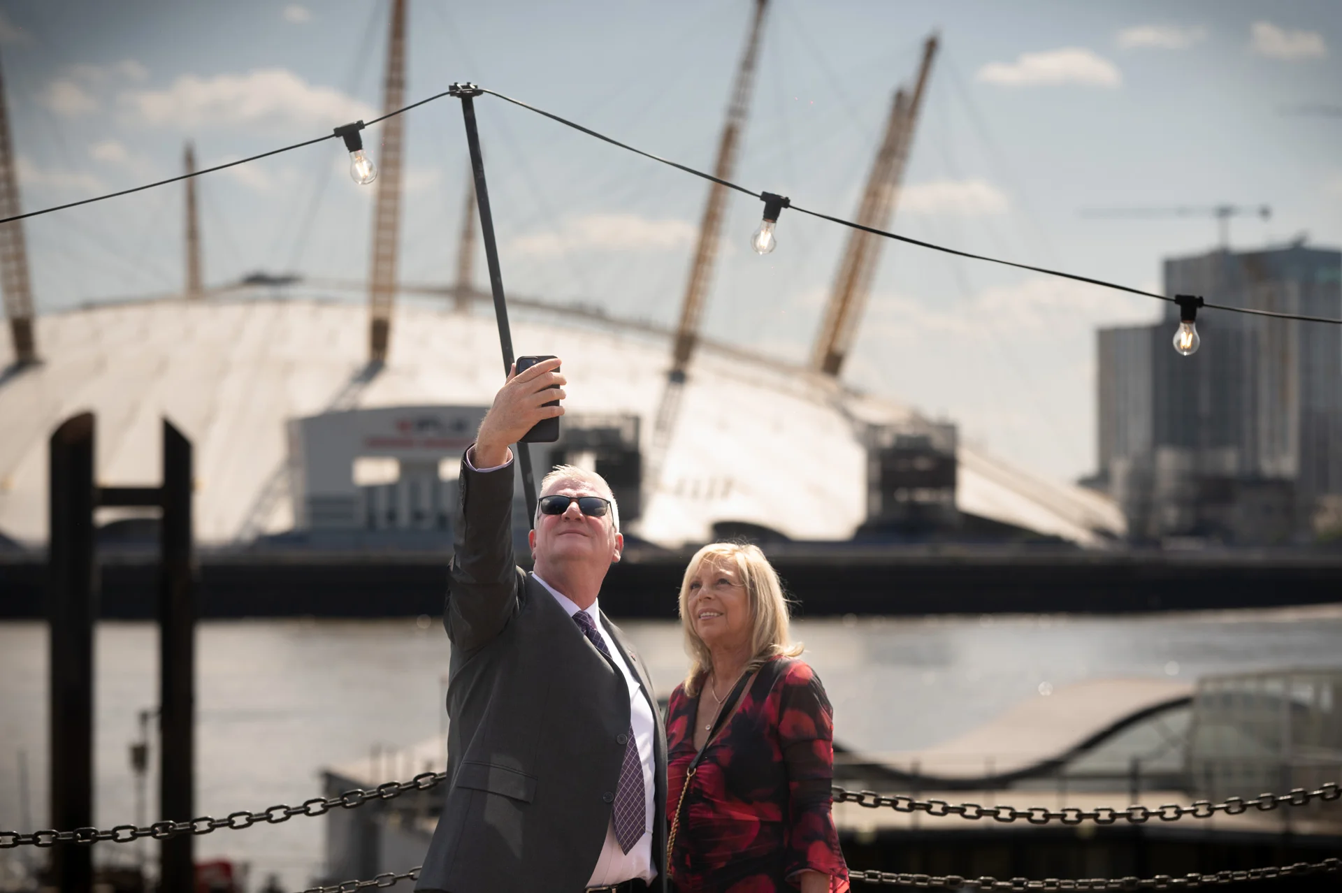 A wedding guest taking a selfie with a woman against a sunny London riverside backdrop featuring the Millennium Dome.
