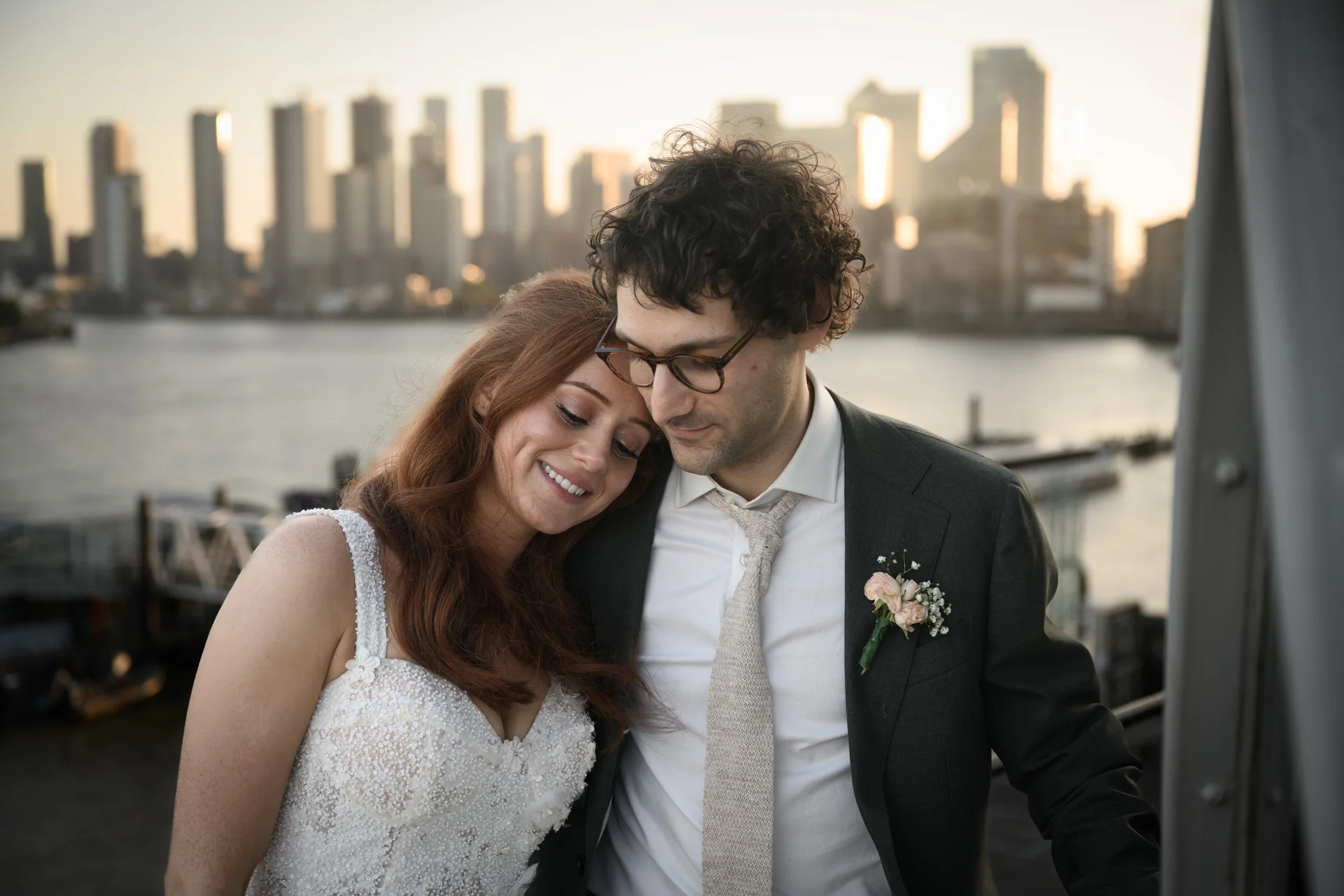 A newlywed couple poses together on a rooftop at sunset, with the bride leaning her head on the groom's shoulder against a striking city skyline backdrop.