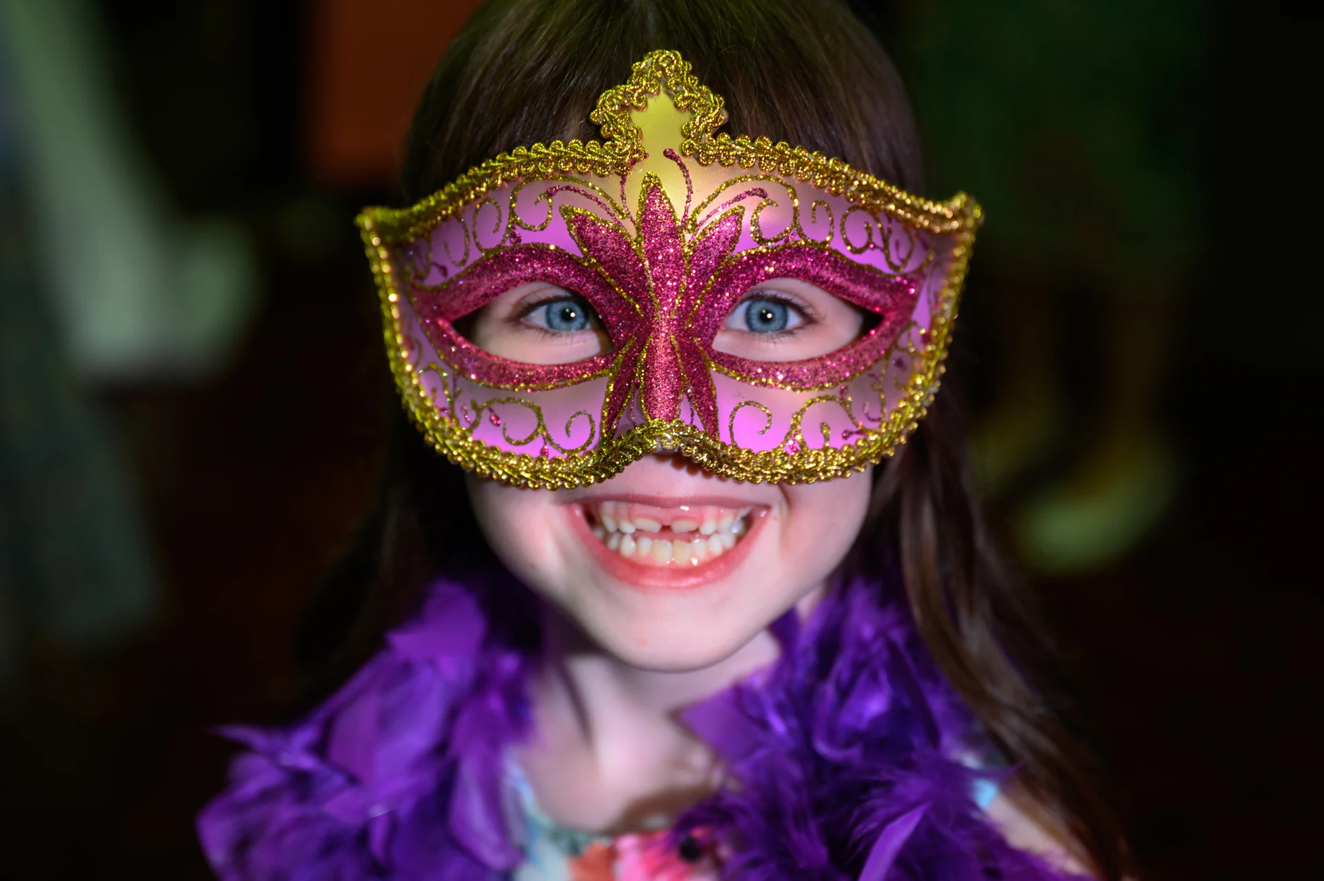 A young flower girl wearing a pink and gold glitter masquerade mask and a purple feather boa smiles joyfully during a wedding celebration.