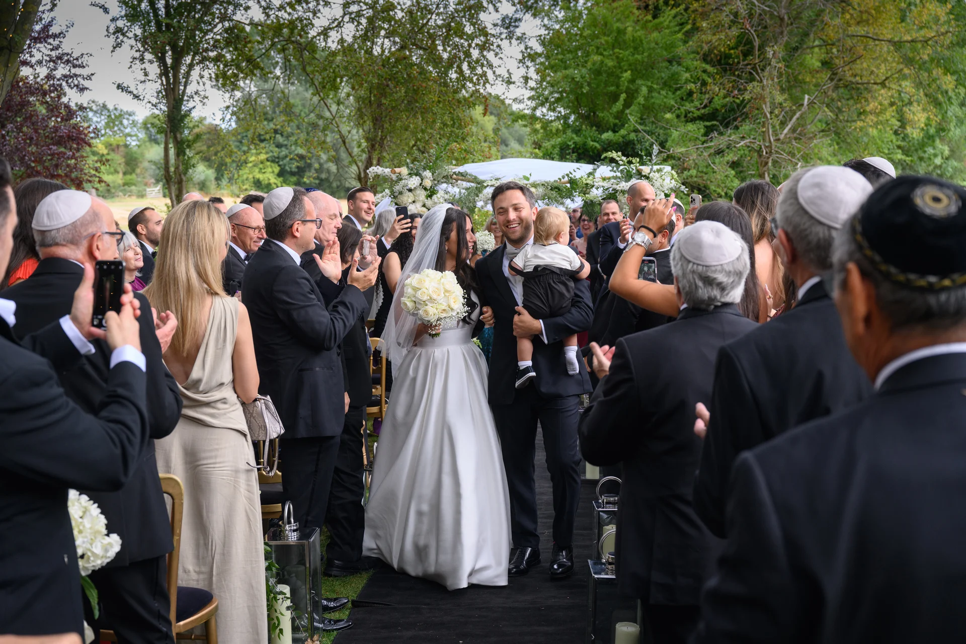 A newlywed couple happily walk back down the aisle together after their outdoor ceremony while guests cheer and applaud, with the groom carrying a young child.