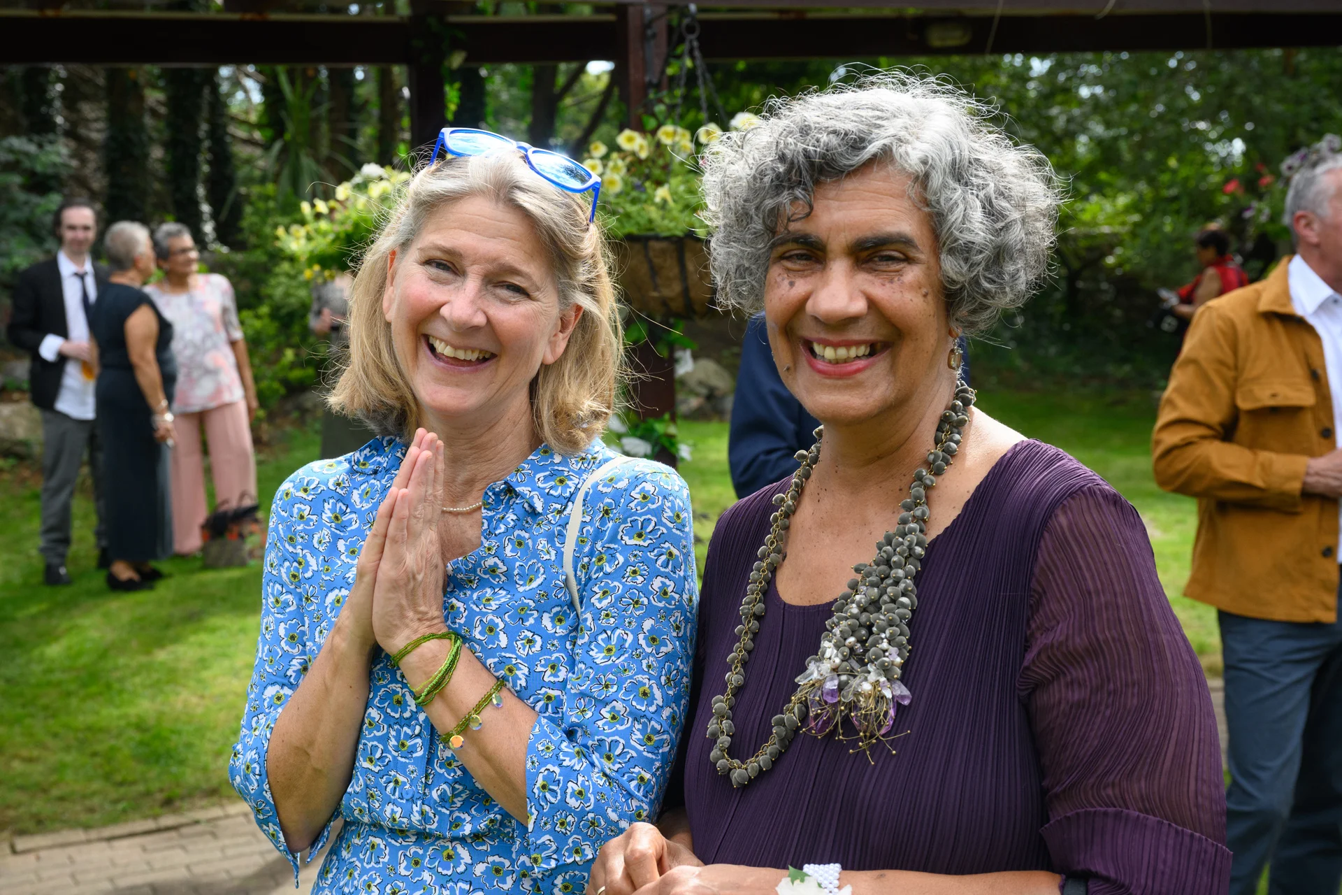 Two smiling guests share a joyful moment outdoors at a wedding reception, surrounded by greenery and other mingling attendees.