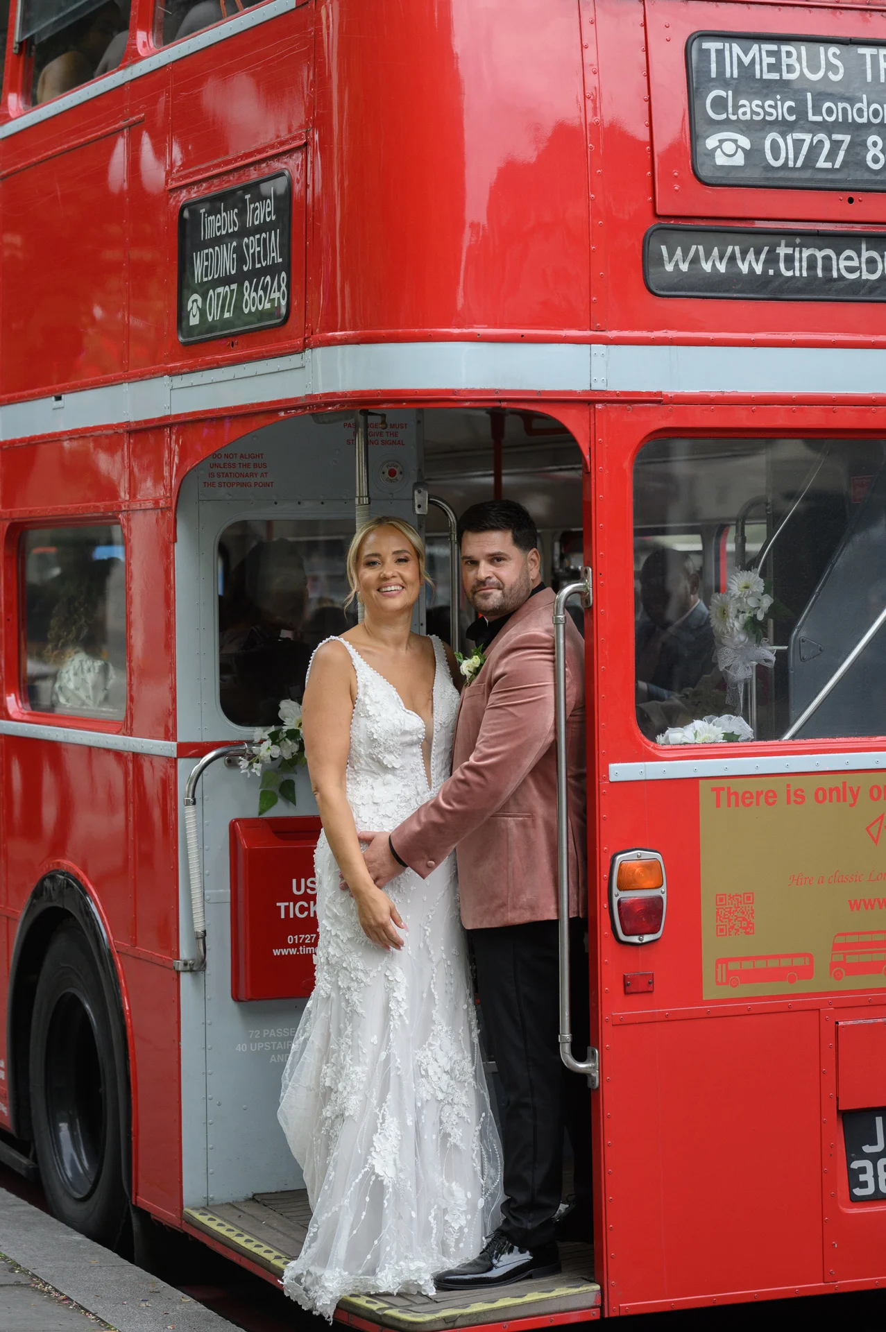 A newlywed couple poses smiling on the platform of a classic red London double-decker bus, with the bride wearing a floral applique gown and the groom in a pink velvet jacket.
