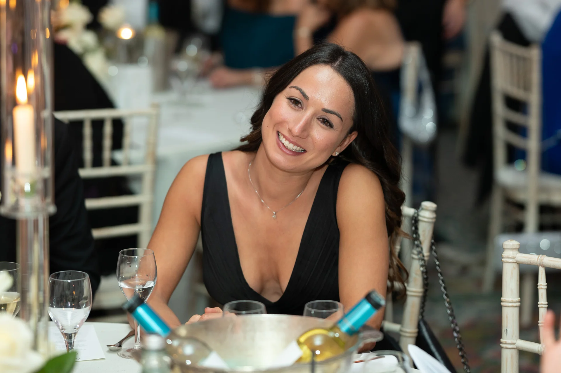 A smiling wedding guest in a black dress enjoys the evening reception seated at a beautifully set dining table.