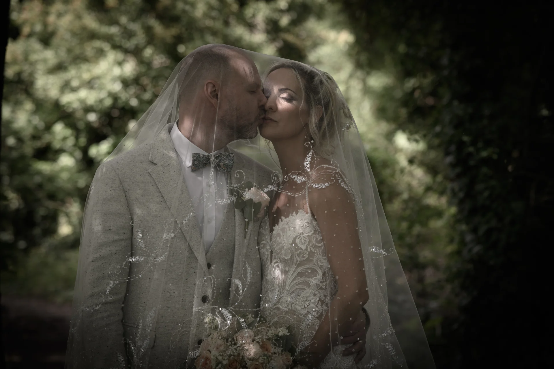A newlywed couple shares a tender kiss beneath an intricately embellished veil in a sun-dappled woodland setting.