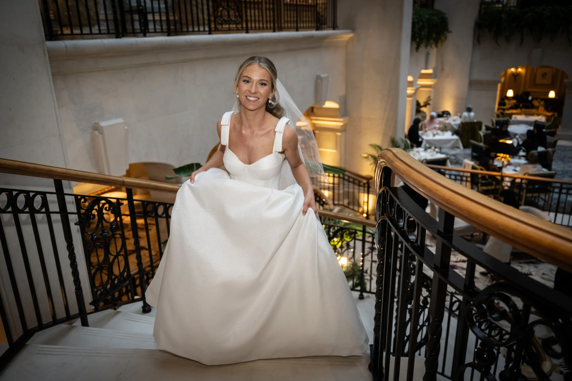 A joyful bride descends a grand staircase inside a luxury hotel, holding up her sparkling satin a line wedding dress.