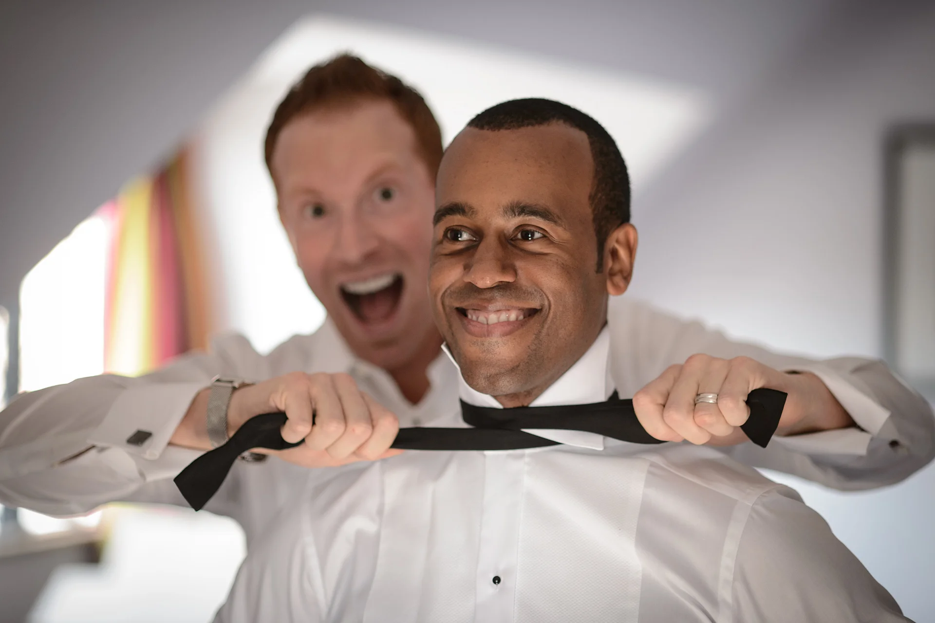 One groom playfully pretends to strangle the other with a black bow tie while both laugh during wedding morning preparations.