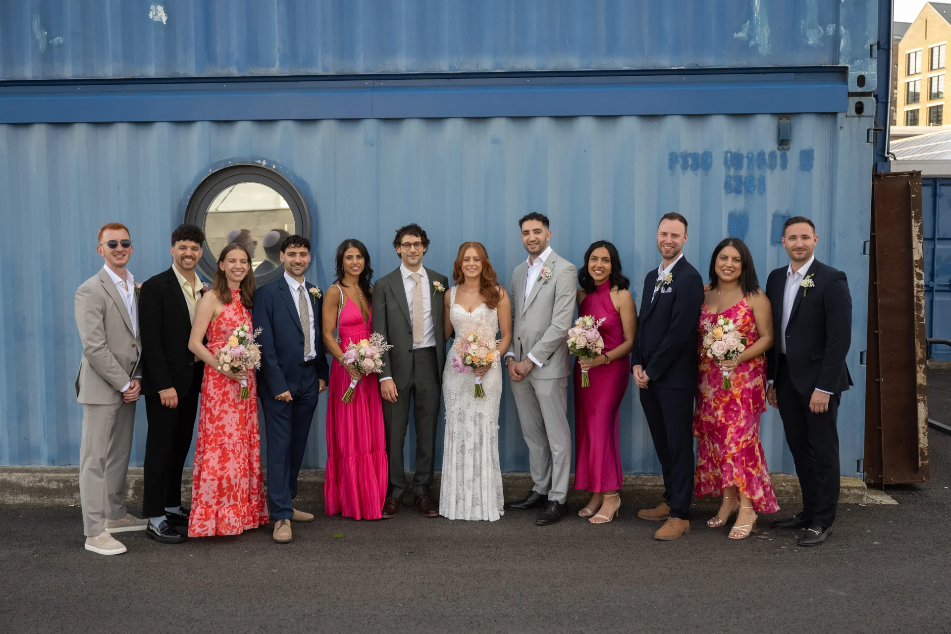 The bride and groom pose alongside a vibrant wedding party in front of a striking blue industrial shipping container wall.