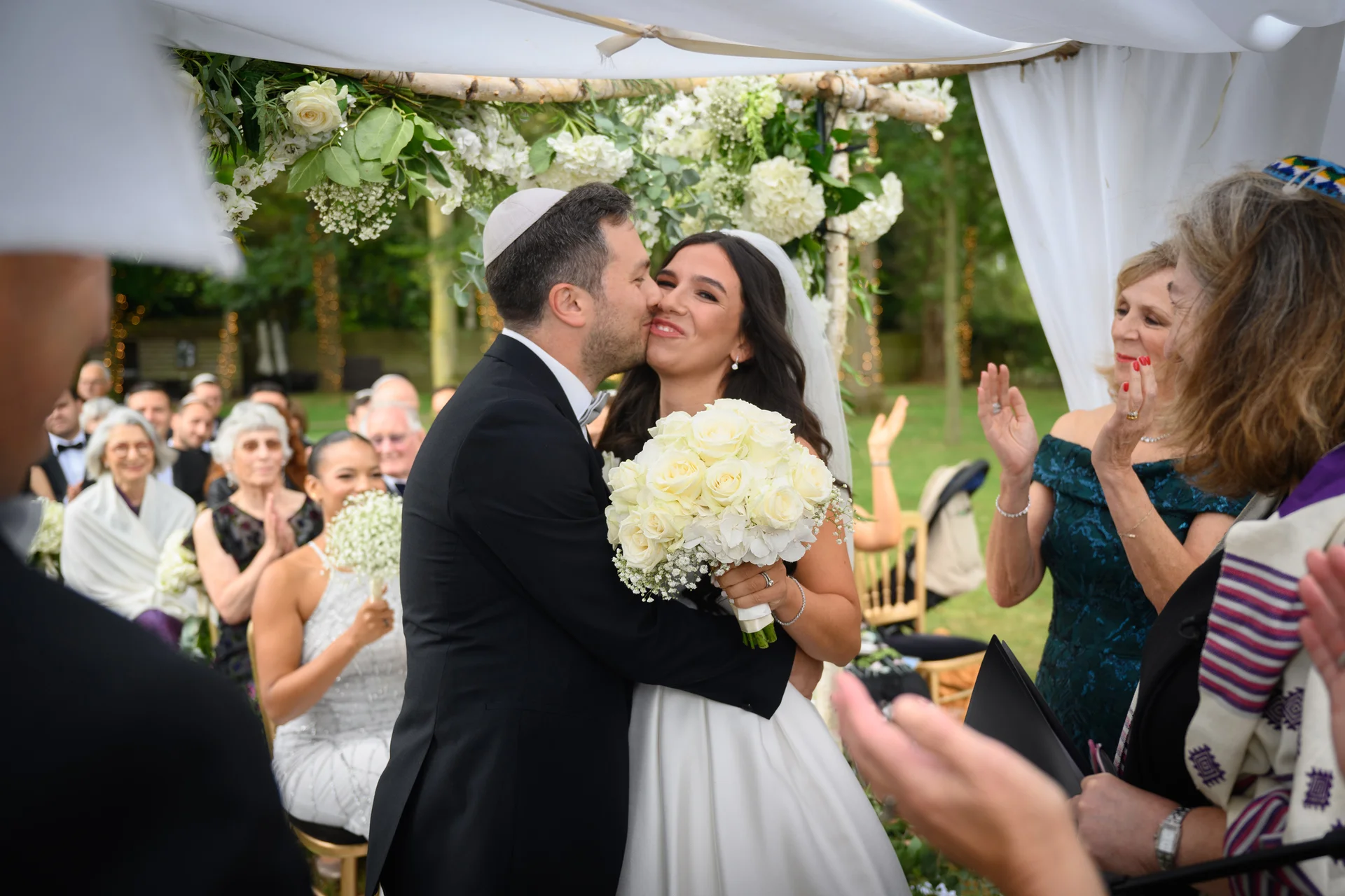 A groom wearing a yarmulke kisses his smiling bride on the cheek beneath a floral chuppah while guests applaud during an outdoor wedding ceremony.