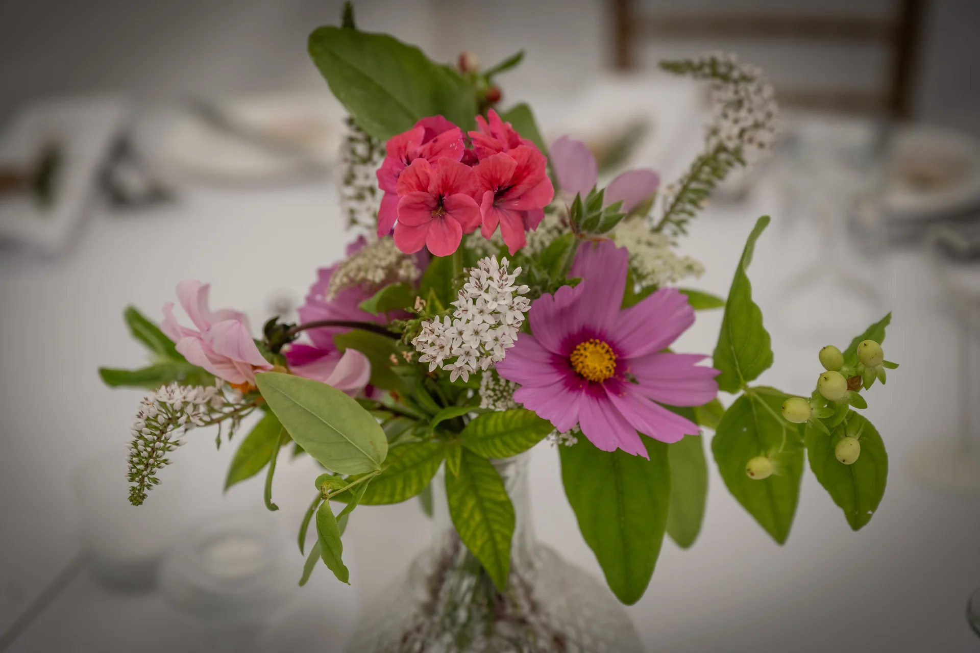 A close-up shot of a small vase filled with a vibrant arrangement of pink cosmos, bright pink geraniums, white filler flowers, and green berries on a wedding reception table.