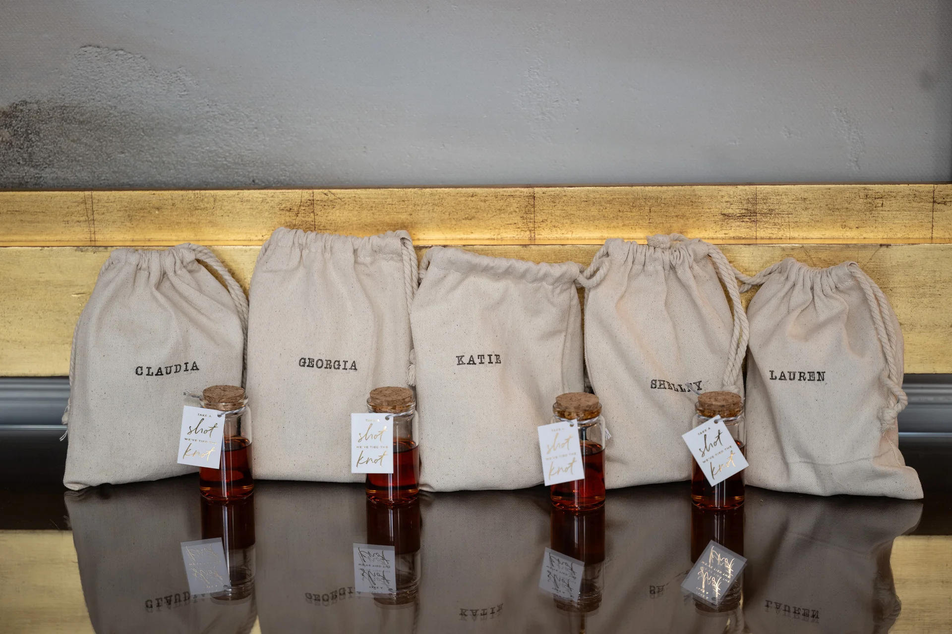 Personalised calico favour bags stamped with guest names are arranged alongside miniature corked bottles of dark liquor on a reflective surface.