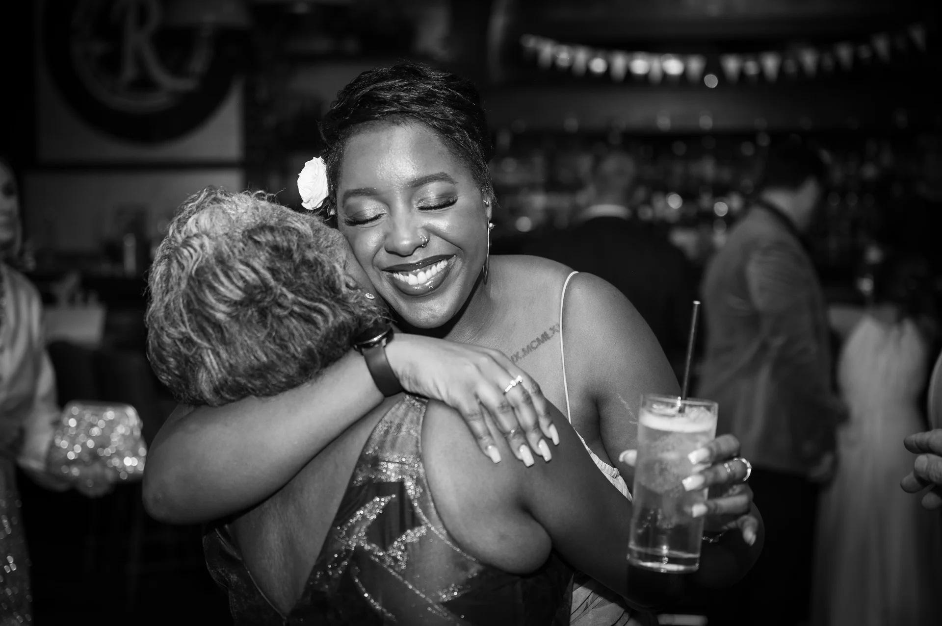 A joyful Black bride with short hair shares a warm, laughing embrace with a guest while holding a drink during the wedding reception.