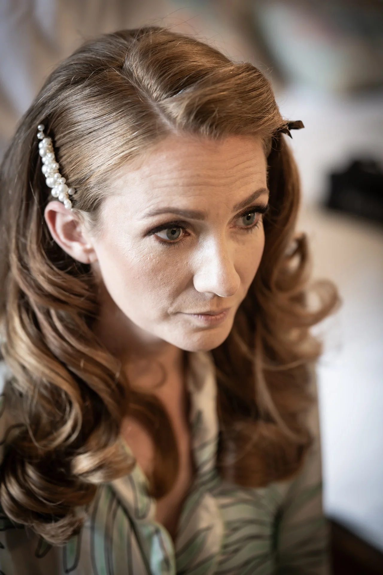 A close-up portrait of a bride getting ready, featuring soft voluminous waves secured with a pearl hair clip and natural dewy makeup.