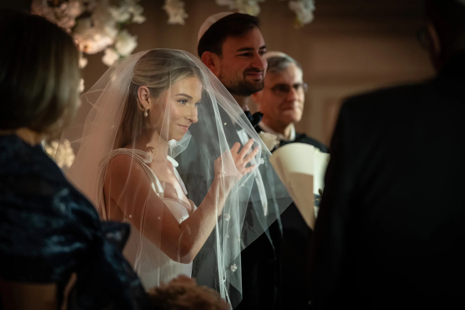 A smiling bride wearing a sheer pearl veil stands beneath the chuppah alongside her groom during a traditional Jewish wedding ceremony.