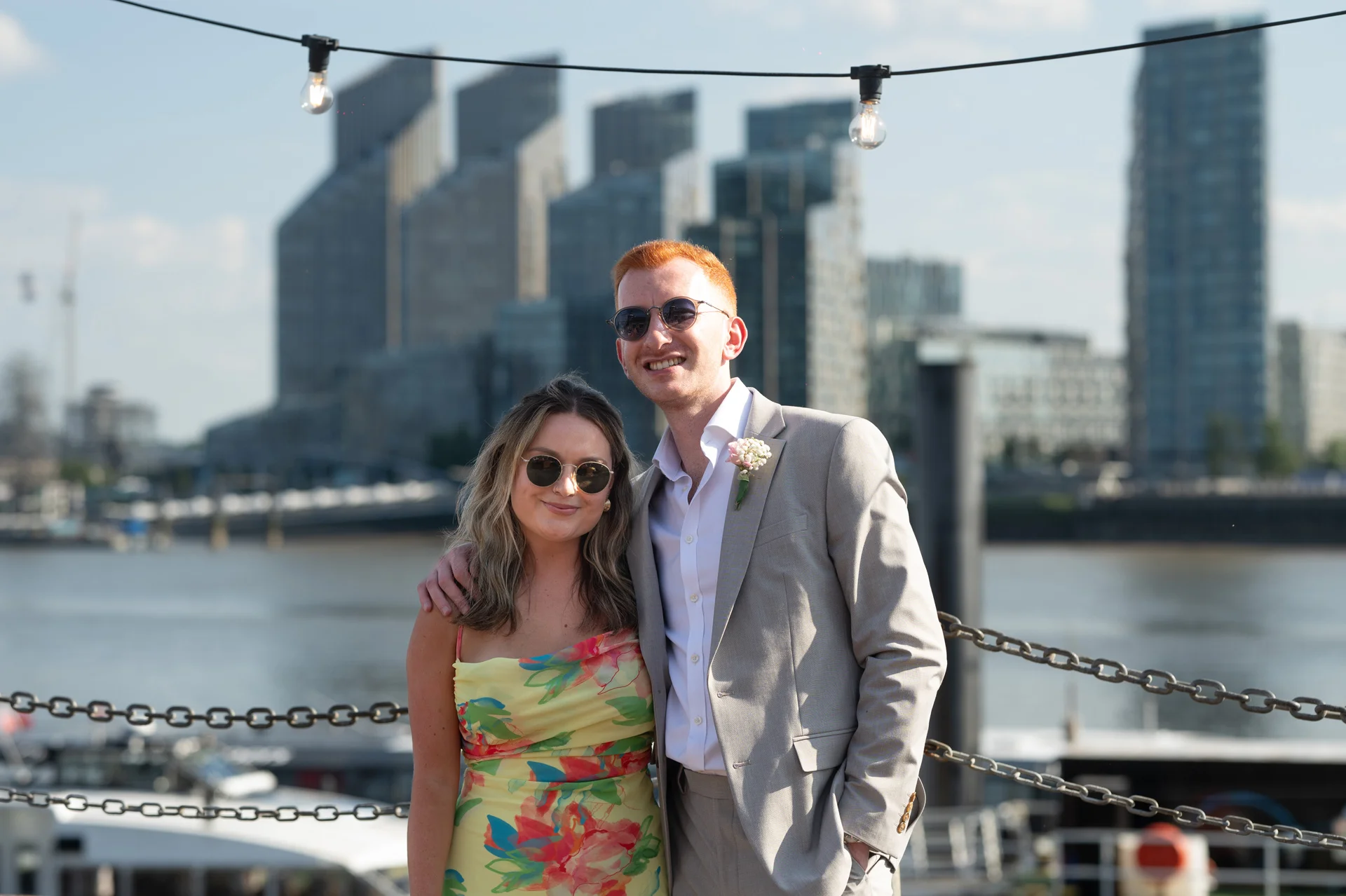 A smiling wedding guest in a vibrant floral dress and a groom in a light grey suit pose outdoors with a modern city skyline and river in the background.