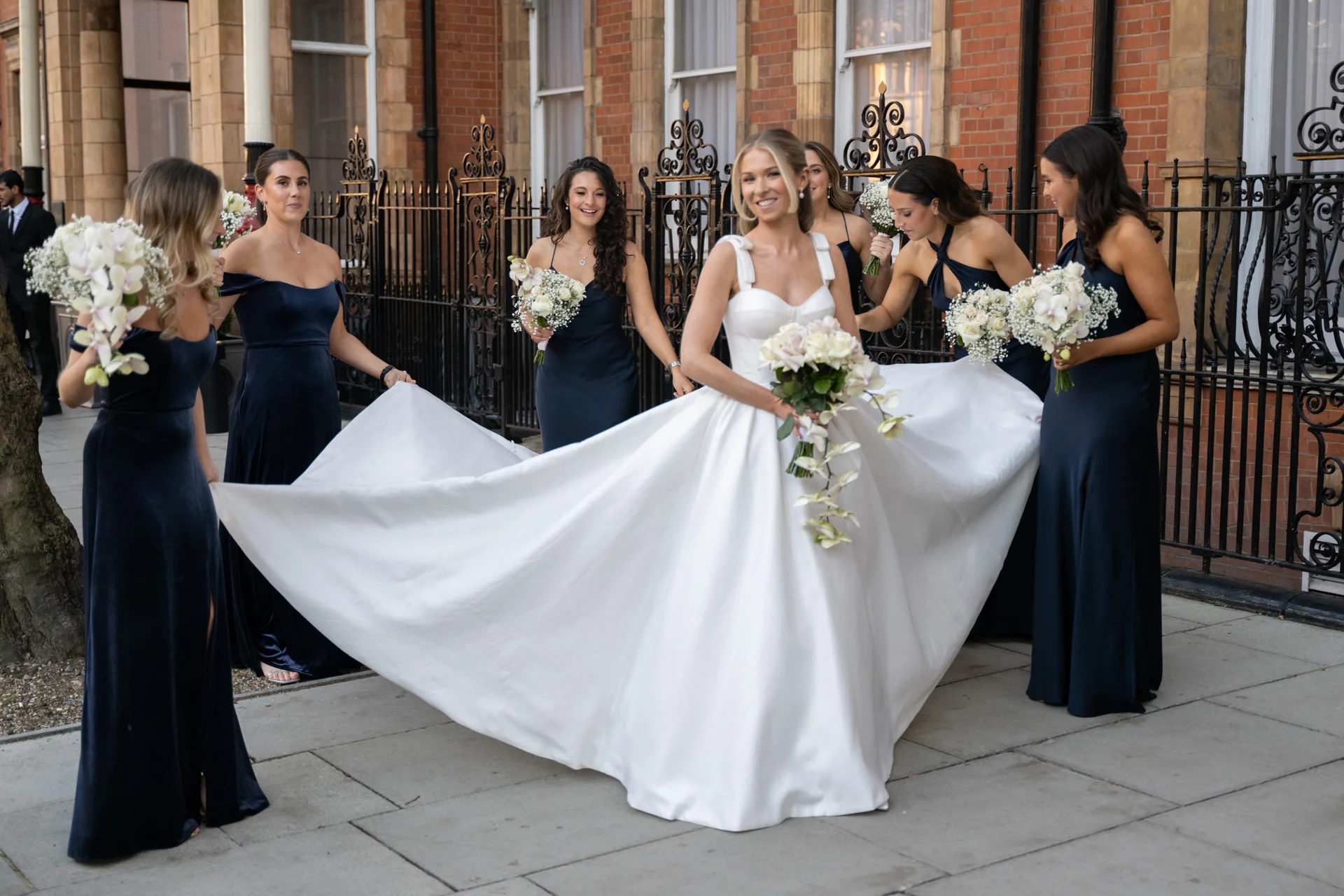 A joyful bride in a satin a-line gown holding a cascading bouquet smiles as her bridesmaids help spread her wide skirt outside a brick building in London.