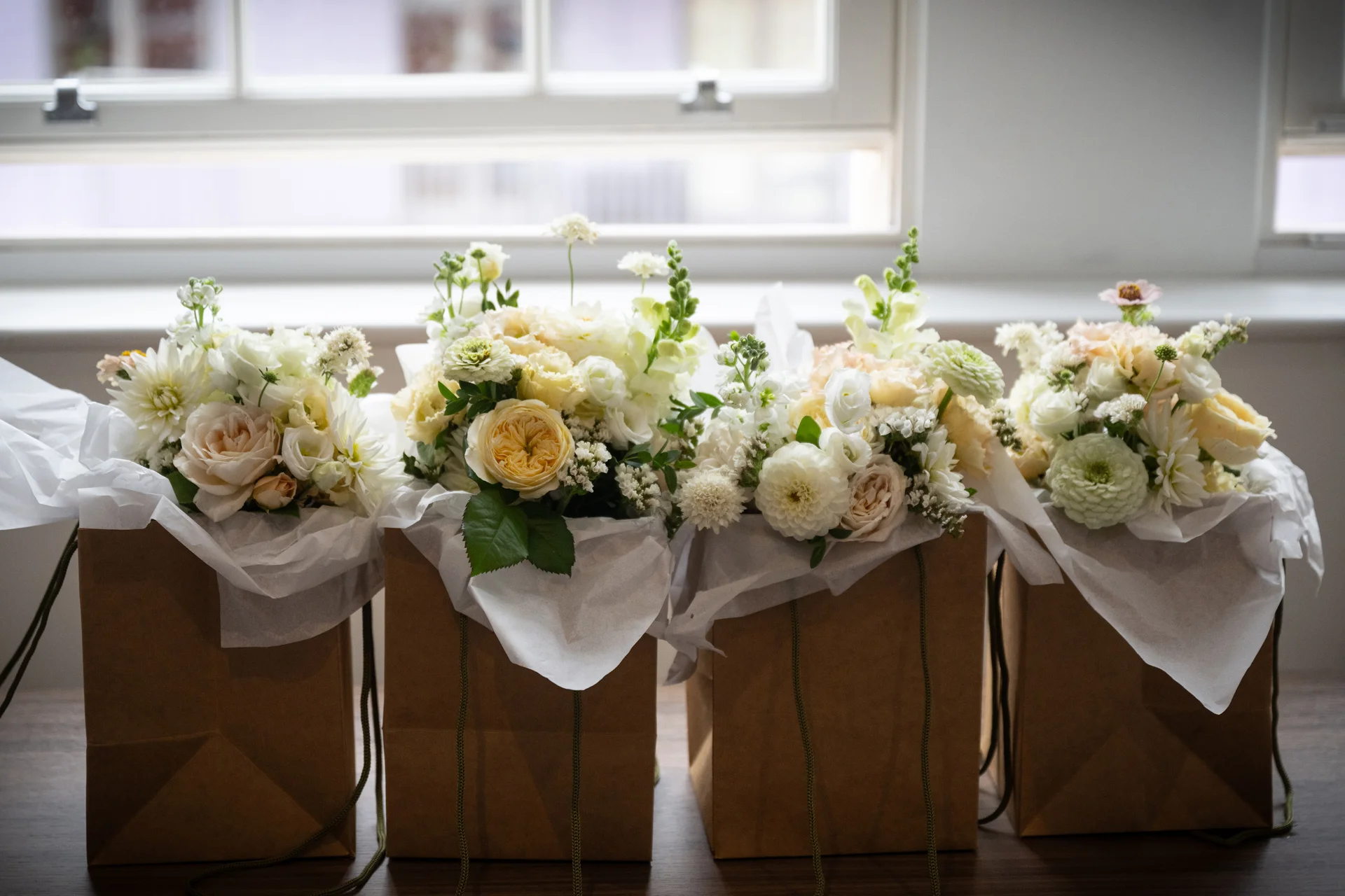 Four kraft paper gift bags filled with delicate tissue paper and lush arrangements of cream and yellow garden roses, ranunculus, and dahlias sit neatly in front of a bright window.