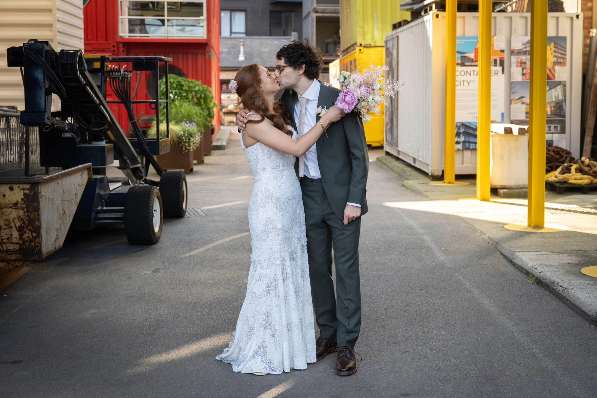 A newly married couple share a tender kiss outdoors in an industrial courtyard setting next to colourful shipping containers.