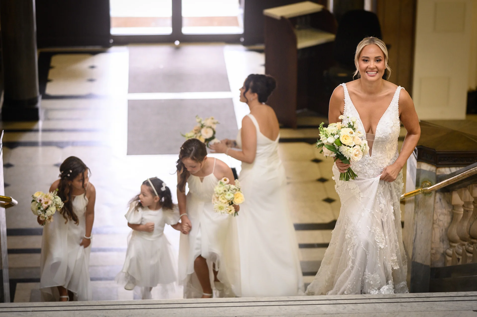 A smiling bride holding a bouquet leads her bridal party and flower girls up a grand stone staircase inside a venue.