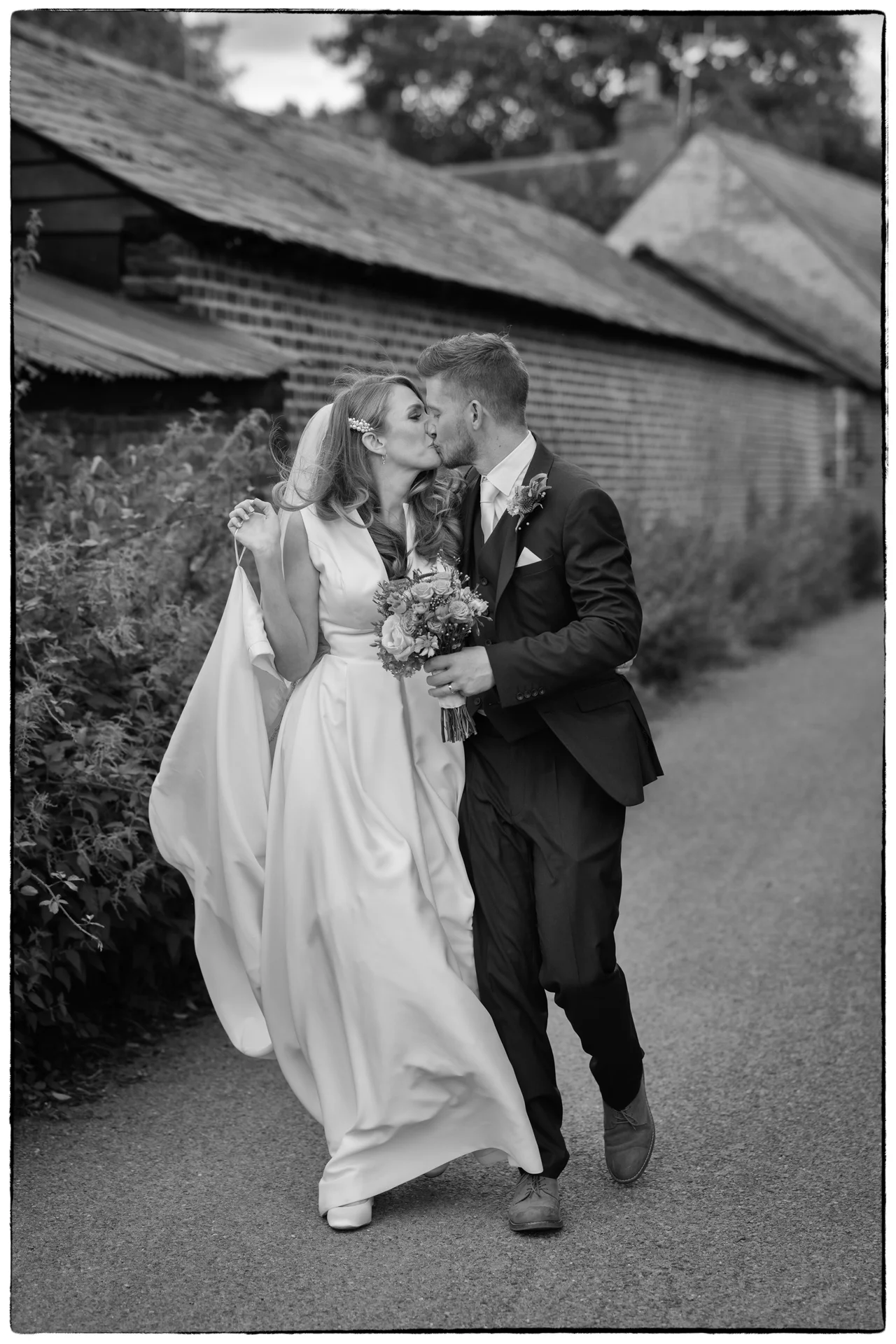 A newlywed couple share a tender kiss while walking together along a rustic country pathway beside old brick farm buildings.