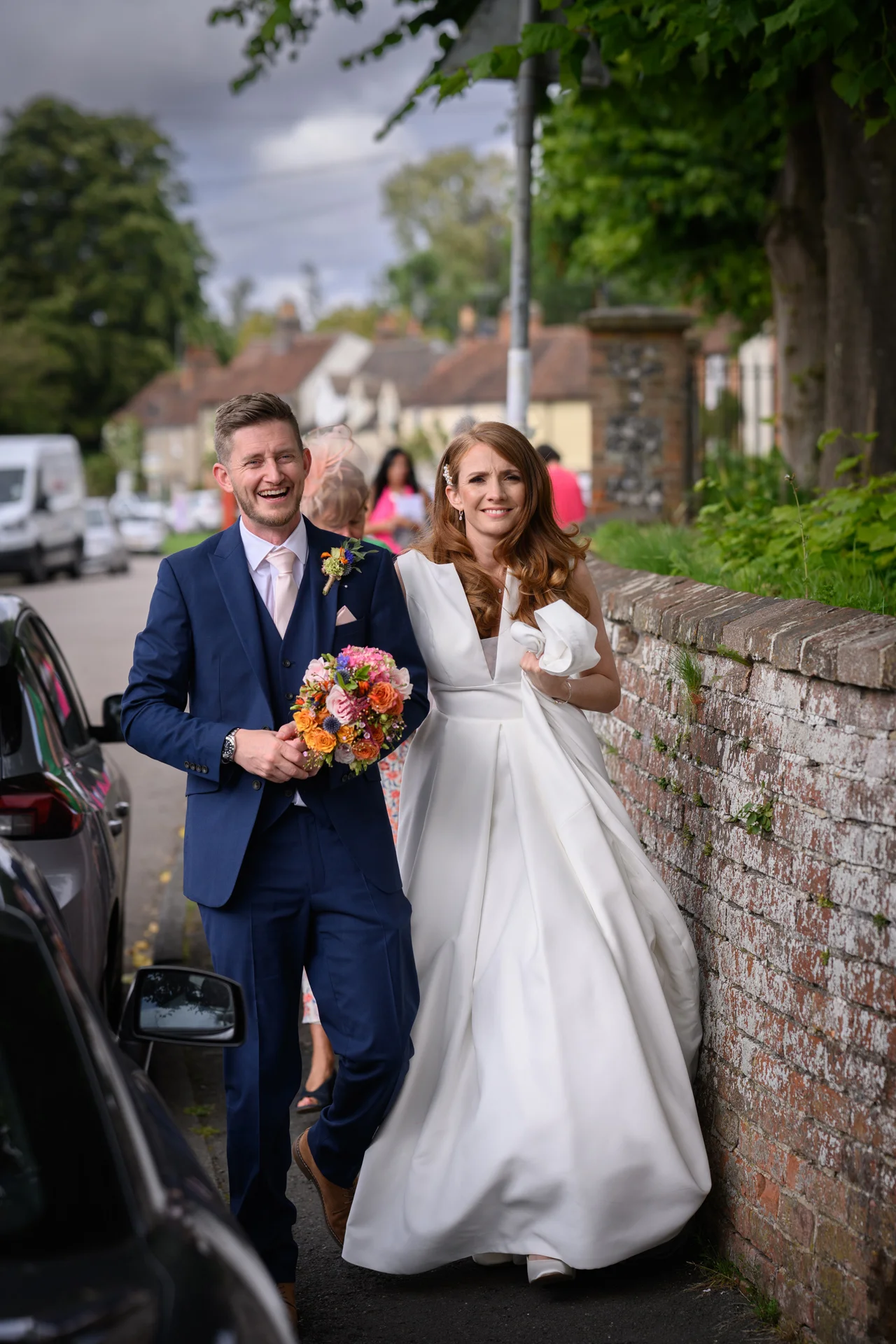 A joyful newlywed couple walking hand in hand along a village street next to a brick wall, with the groom laughing heartily in a navy suit and the bride smiling warmly in a white satin a-line gown while holding a colourful bouquet.