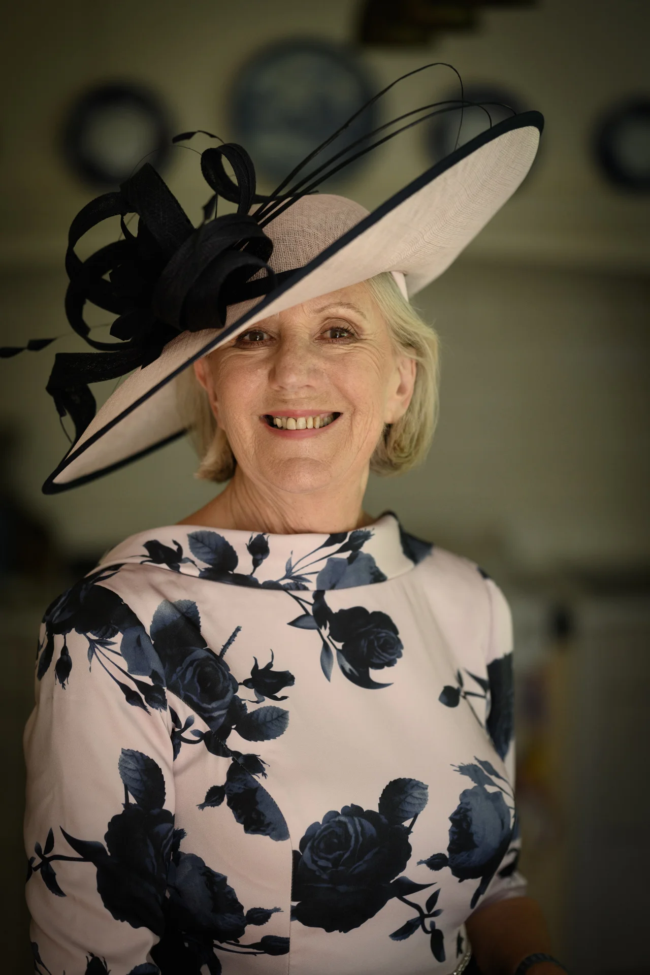 An elegant mother of the bride smiles warmly wearing a wide-brimmed pink hat with black trim and a matching floral print dress.