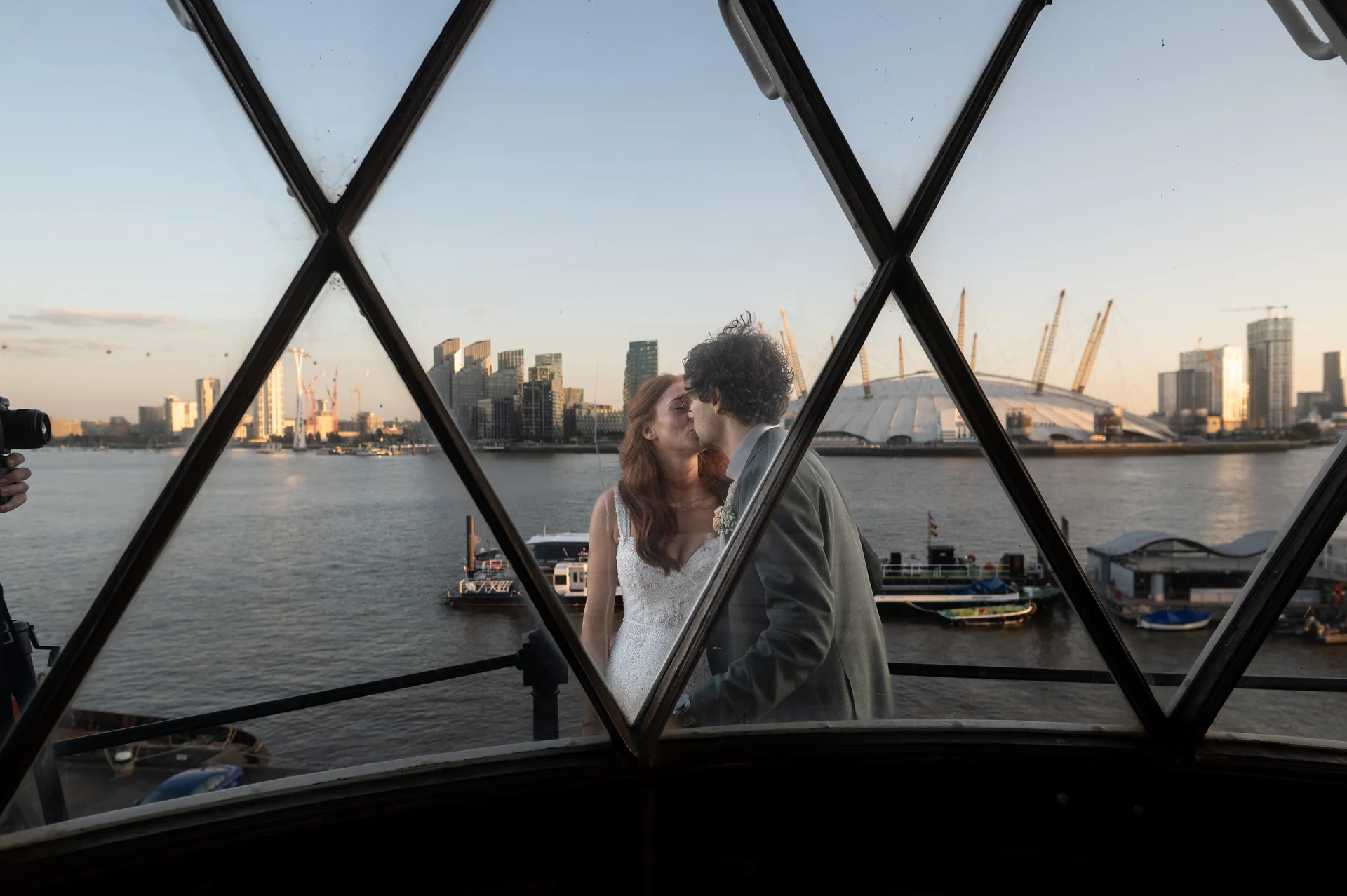 A newlywed couple shares a tender kiss by the riverside, viewed through a patterned window overlooking a London city skyline at sunset.
