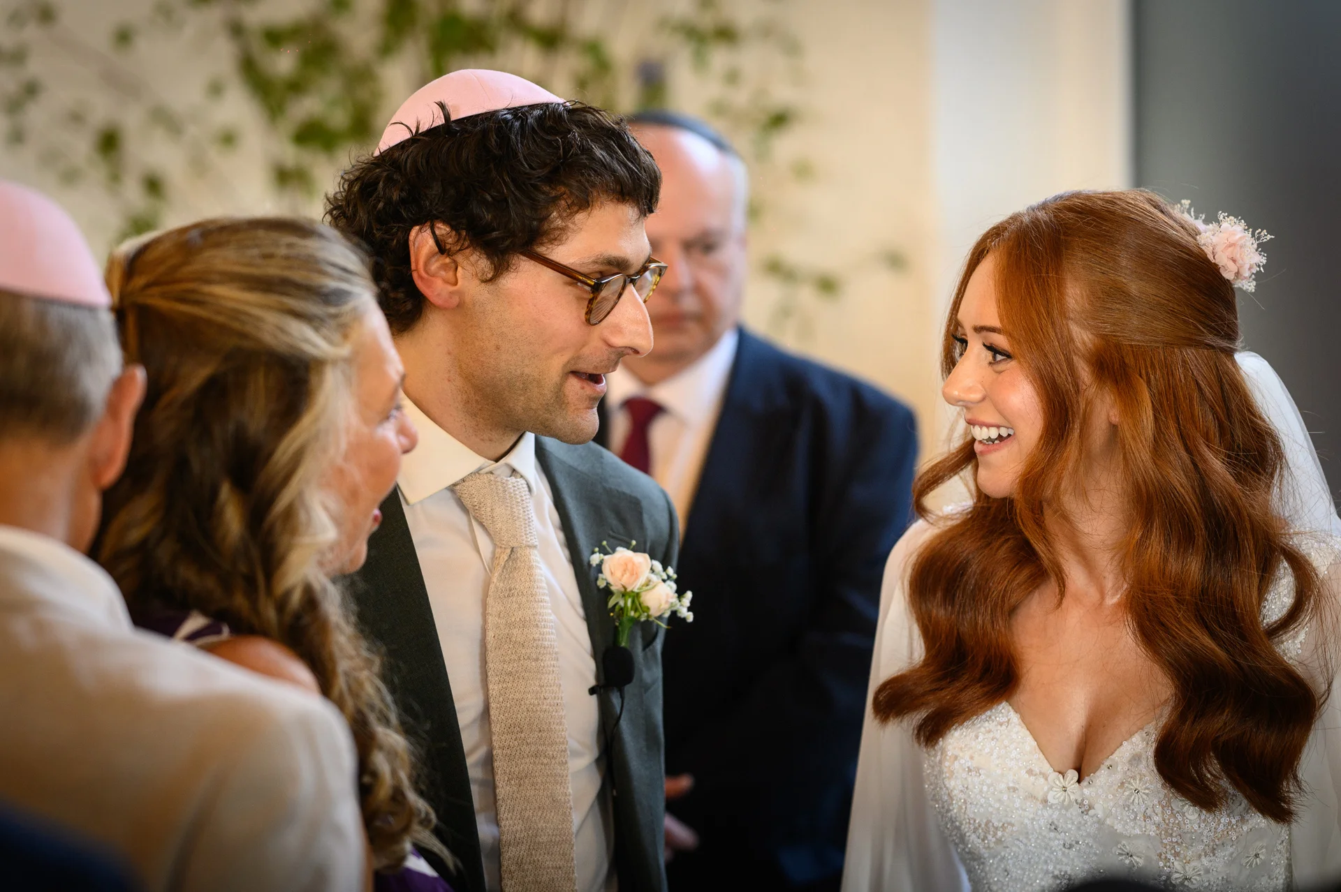 A joyful bride and groom smile at each other during a Jewish wedding ceremony, surrounded by family members wearing kippahs.