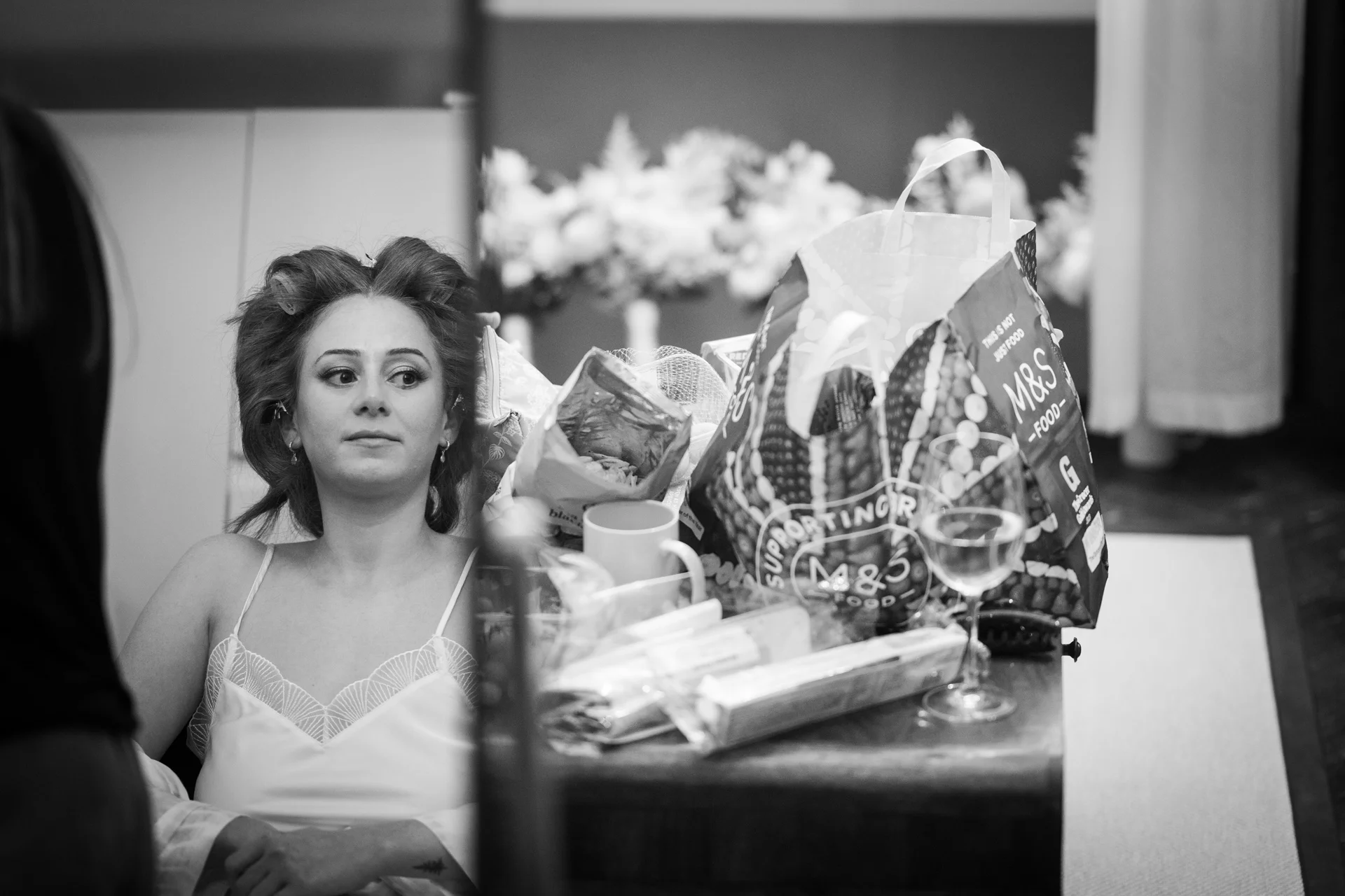 A black and white photograph captures a bride sitting in front of a mirror with hair rollers during morning preparations while a shopping bag and snacks sit nearby.