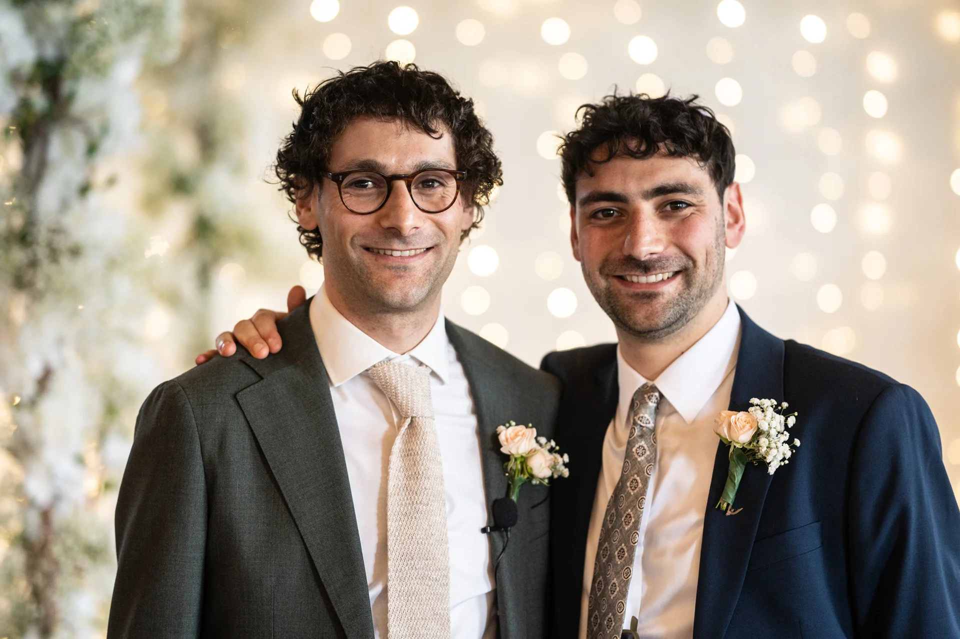 Two smiling grooms in tailored suits with buttonholes pose together in front of a softly lit fairy light backdrop.