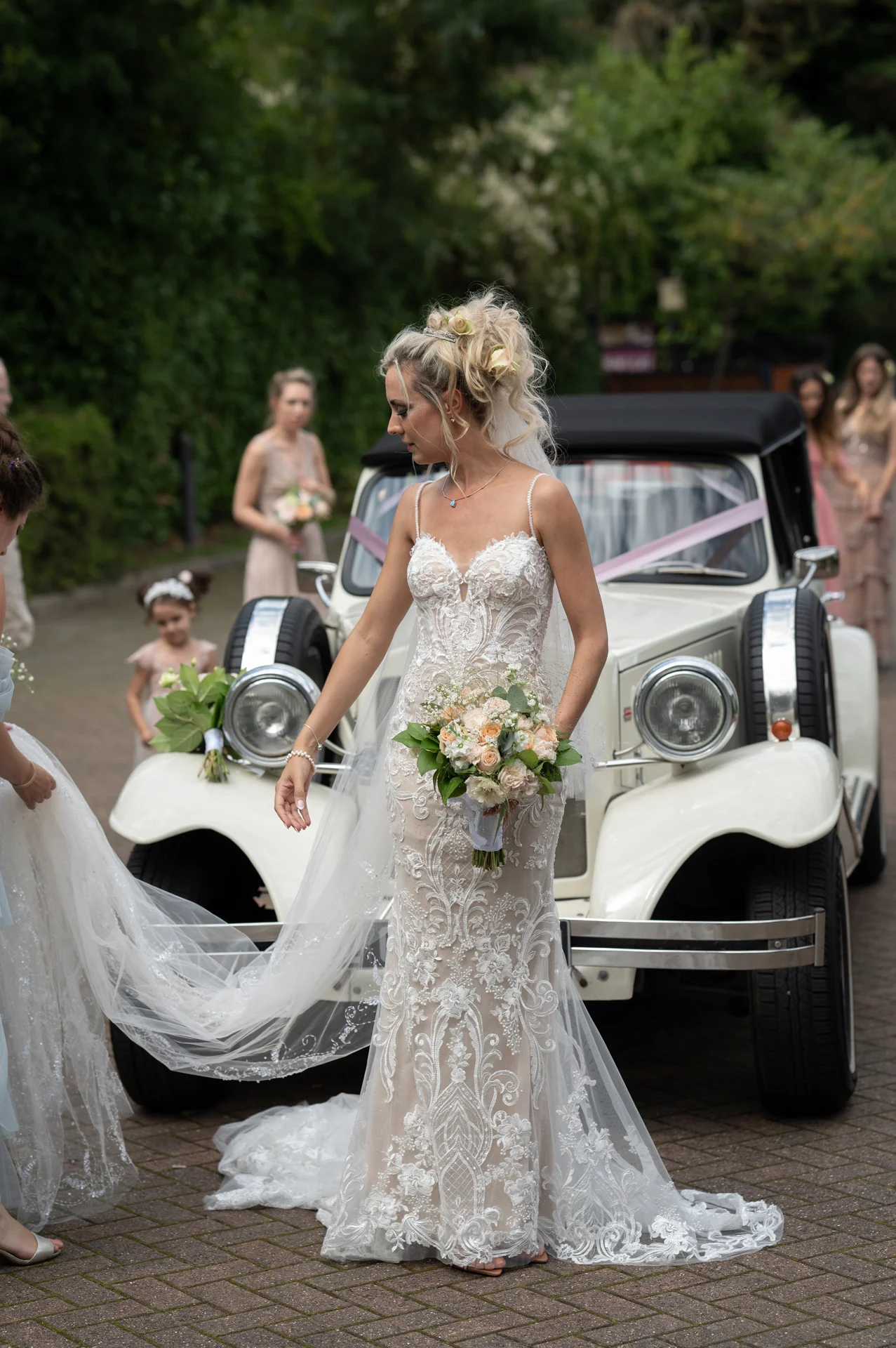 A stunning bride in an embroidered lace sheath dress stands beside a cream vintage wedding car, holding a pastel bouquet while someone helps arrange her veil.